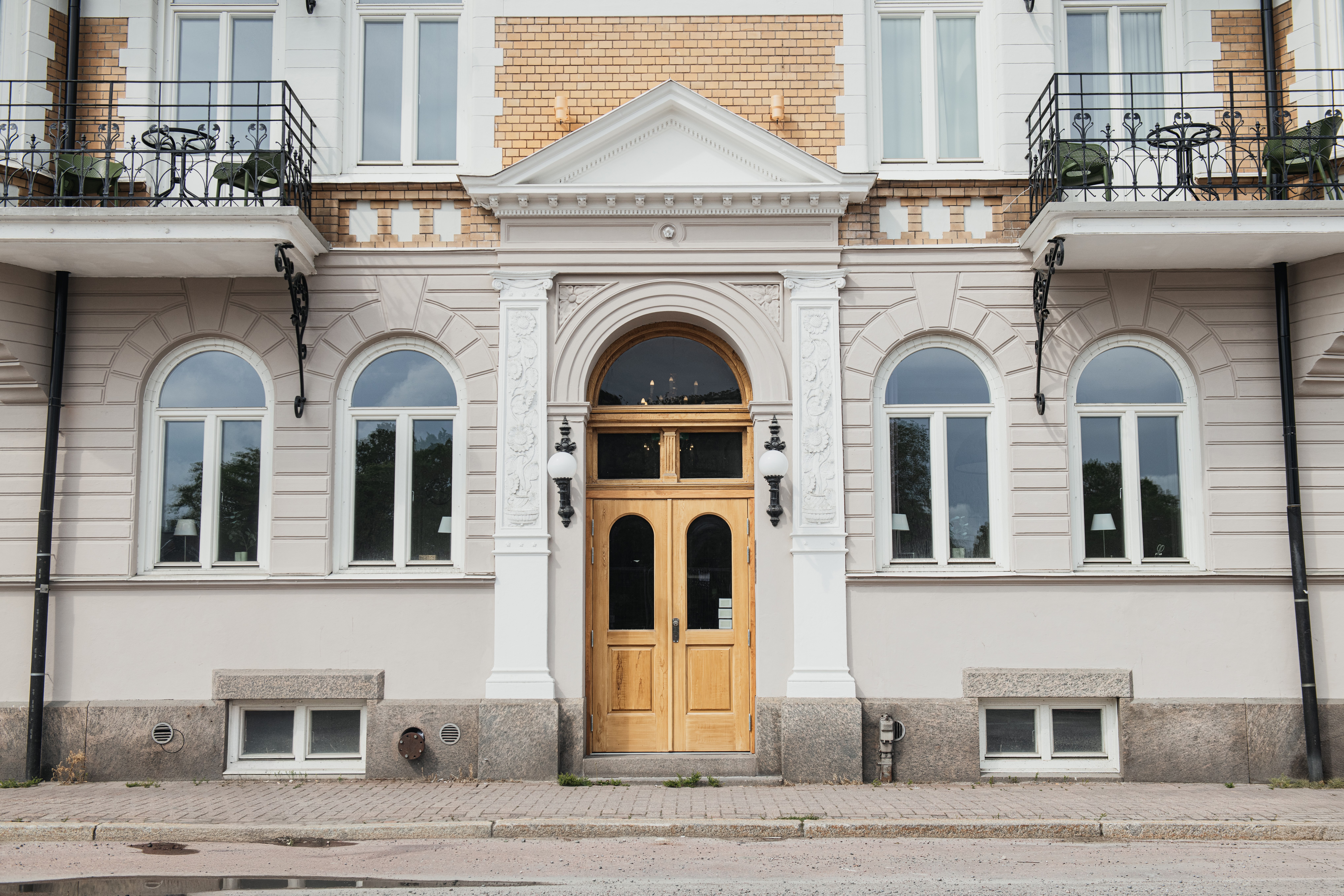 Ett par trädörrar sitter i centrum av en klassisk fasad med välvda fönster och dekorativa kolumner. Tegel- och putsbyggnaden har små balkonger och stensteg till ingången.