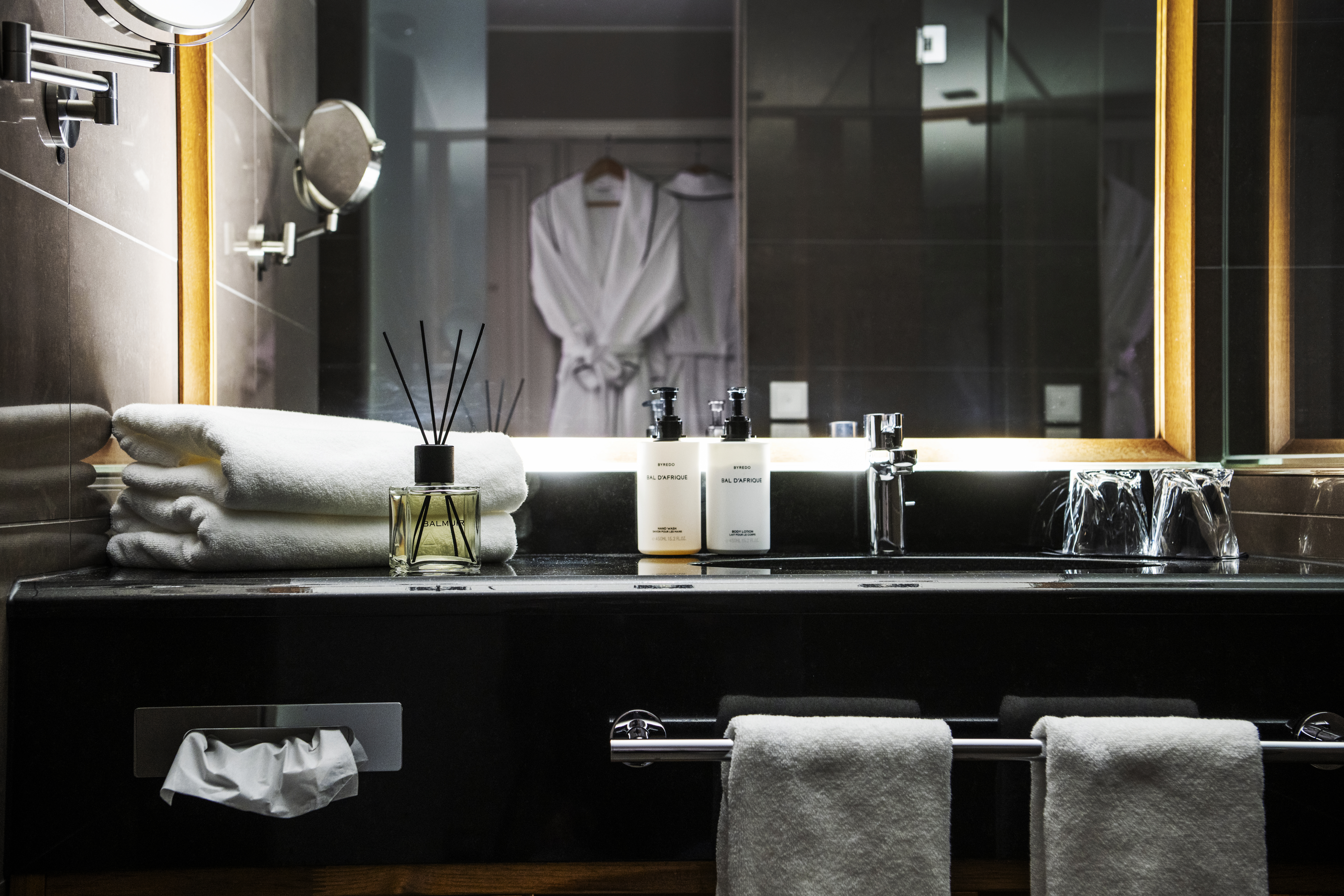 A bathroom counter holds stacked white towels, toiletries, and a reed diffuser. A mirror reflects two hanging robes, illuminated by warm lighting. 