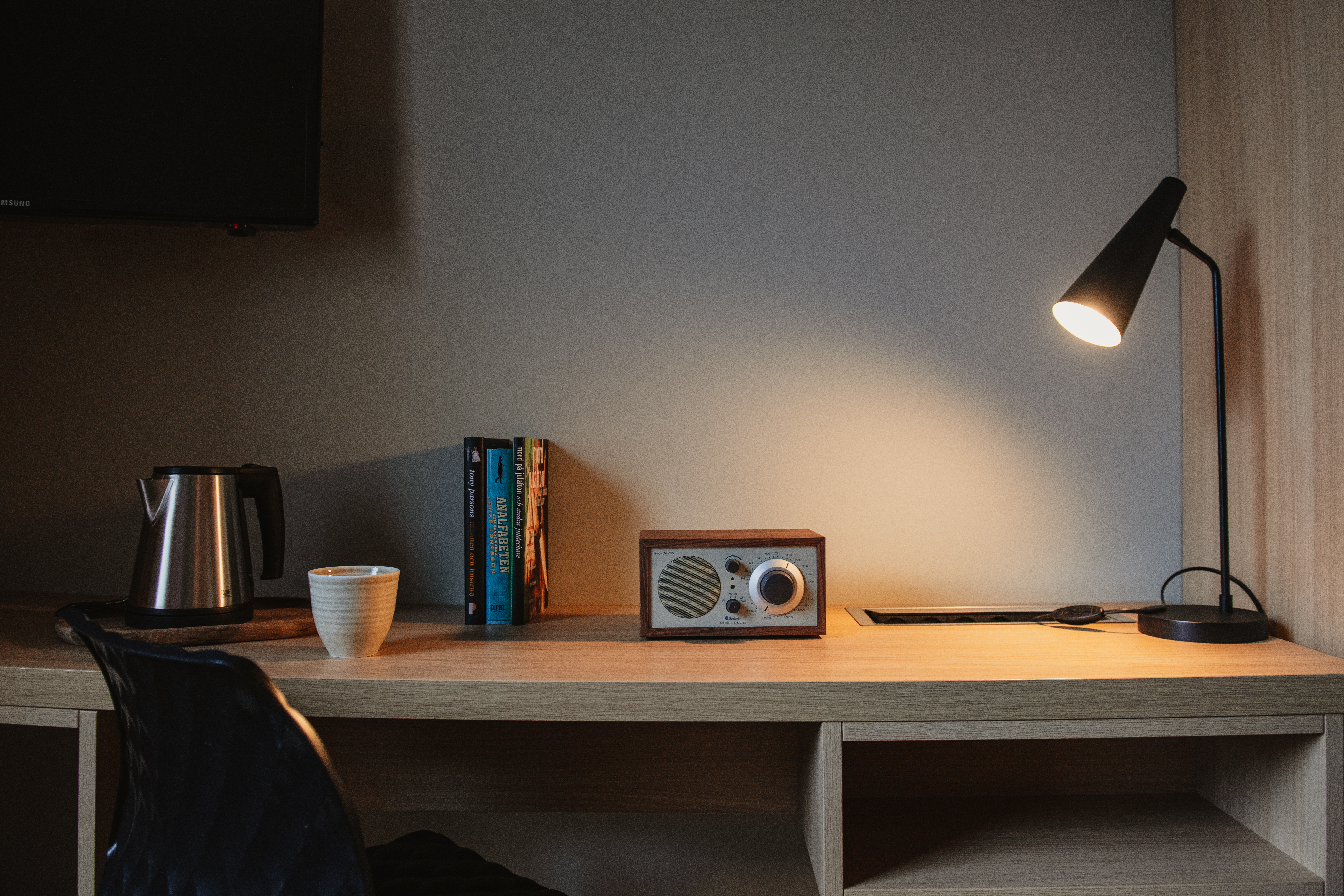 Ett träskrivbord har en retro radio bredvid en tänd svart lampa. I närheten står en vattenkokare och en mugg, med böcker prydligt uppradade i en modern kontorsmiljö.
