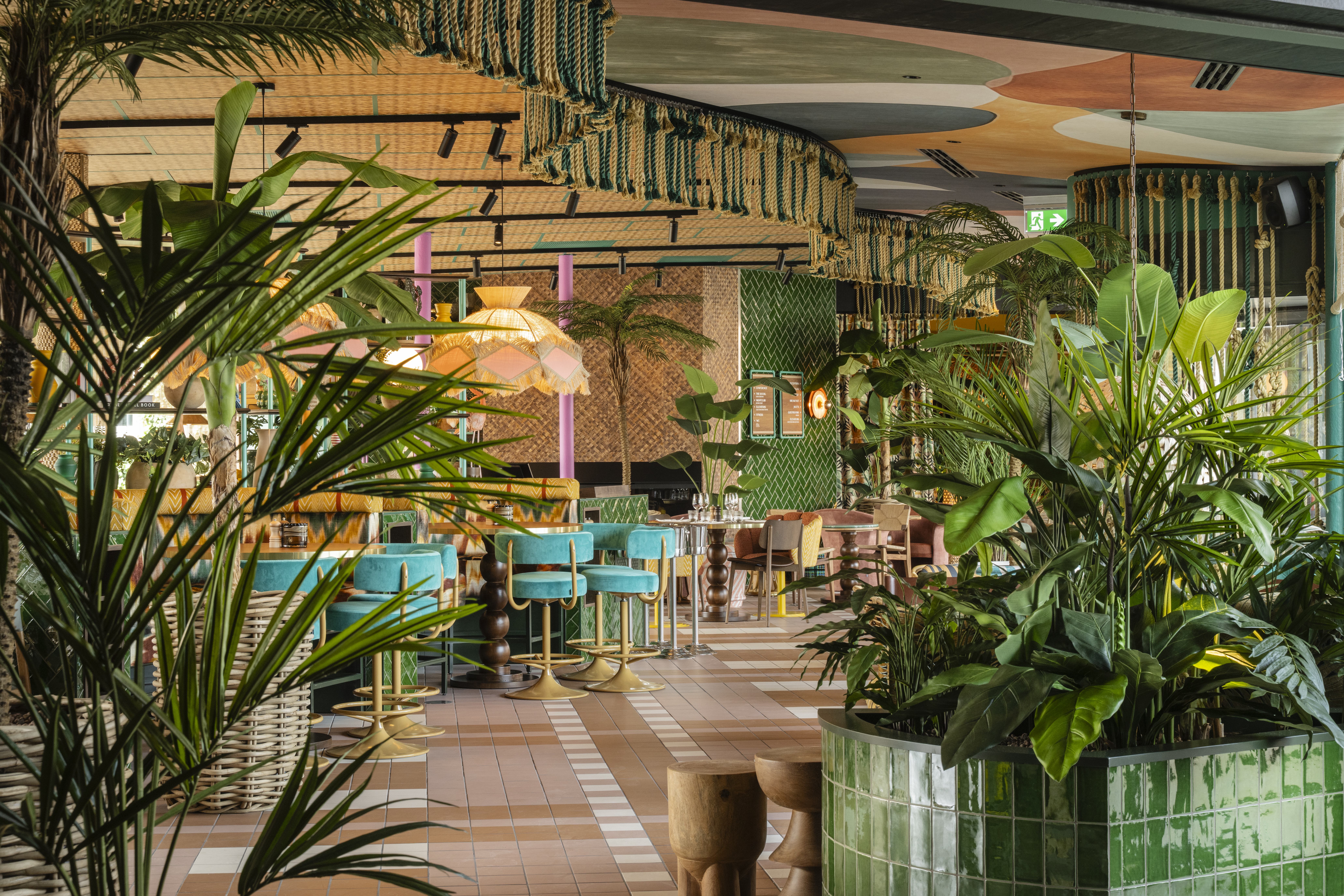 Frodig vegetation fylder en restaurant med farverige siddepladser og hængende stofdekorationer. Grønne fliser, livlige møbler og tropiske planter skaber en livlig, indbydende atmosfære.