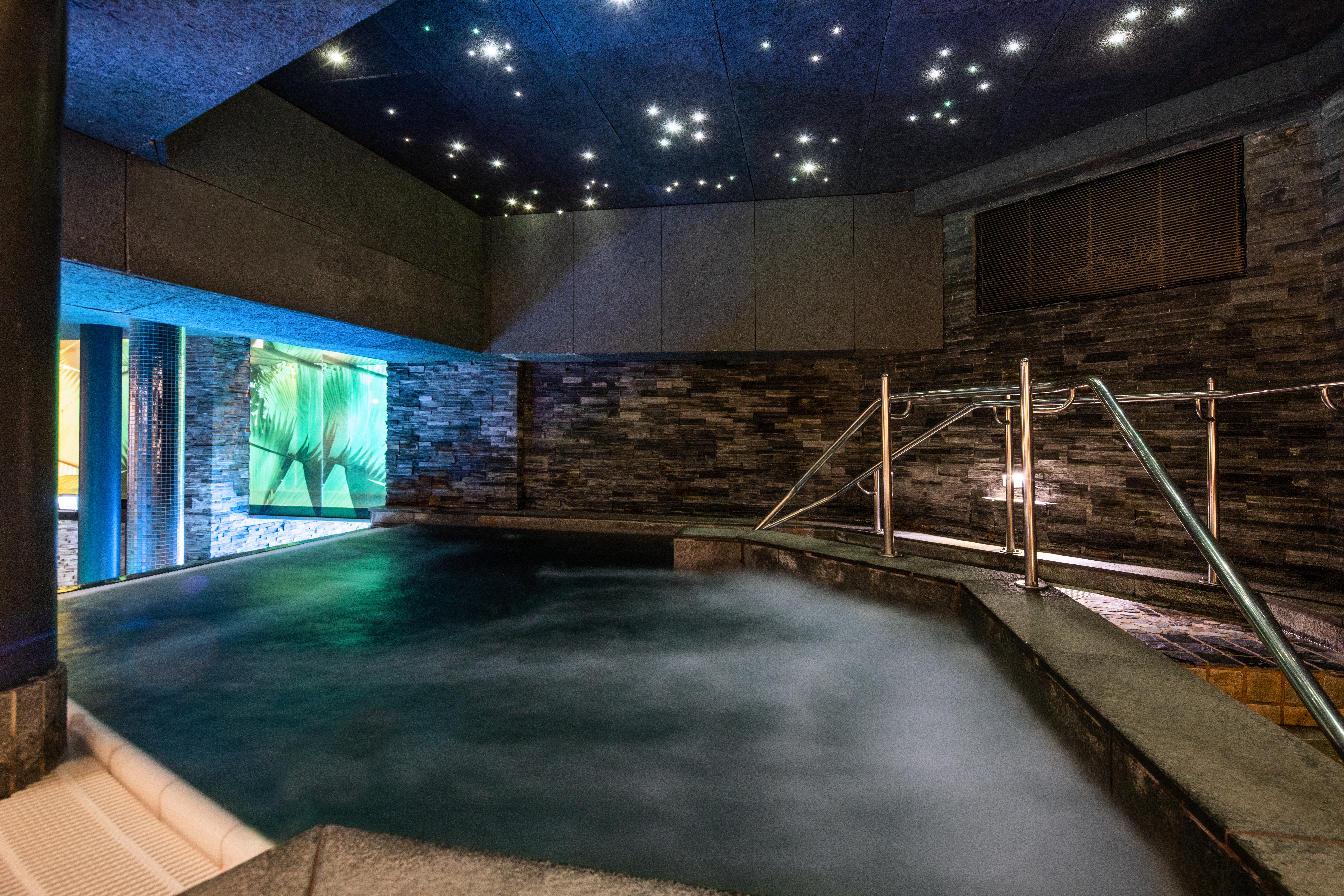 En rolig indendørs pool med let damp, omgivet af stenvægge. Et metalgelænder fører ned i vandet, under et loft med stjernelignende lys.