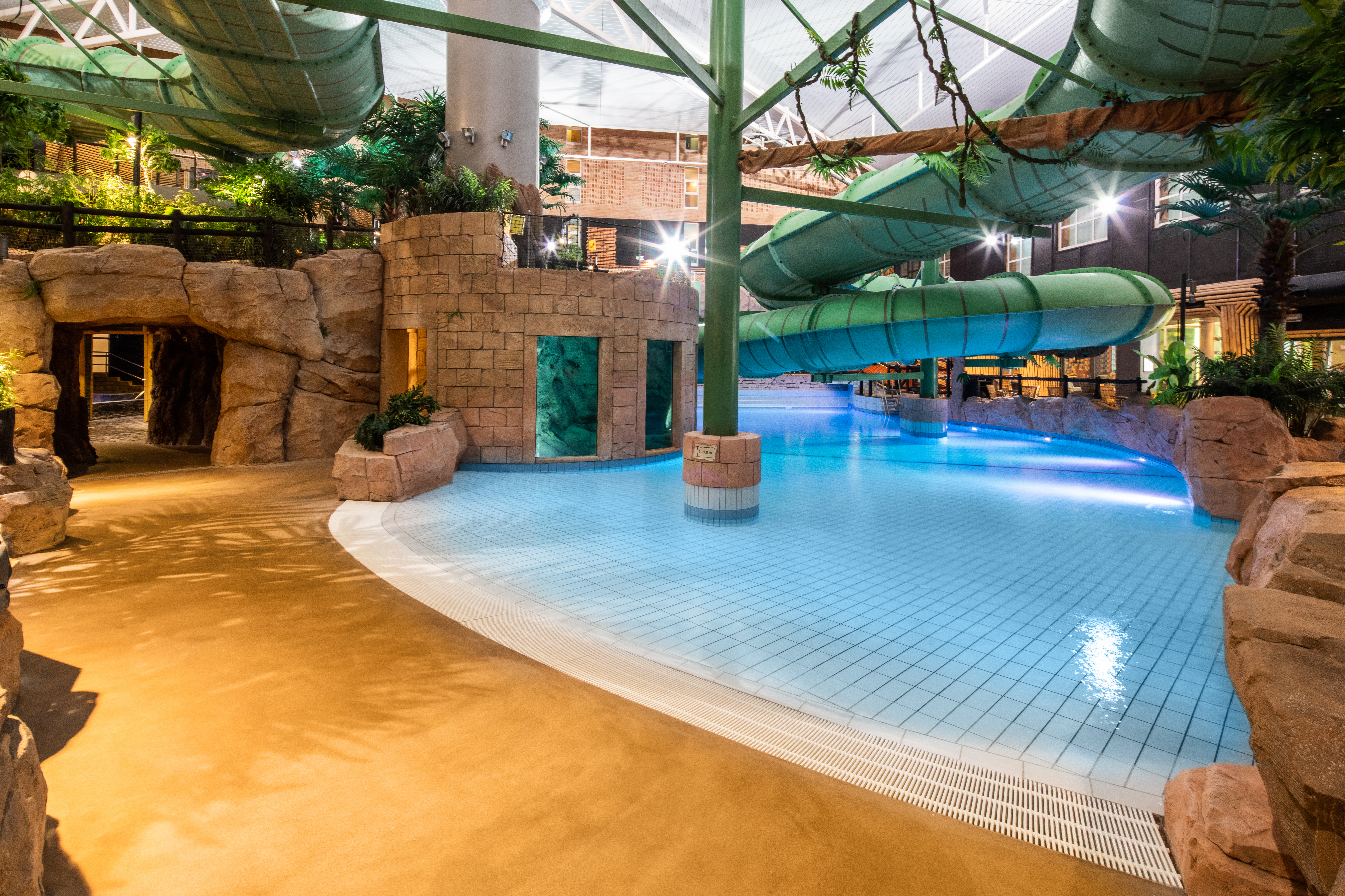 En stor grøn vandrutsjebane bøjer sig over en klarblå pool i en indendørs vandpark. Området er omgivet af kunstige klippeformationer og planter.