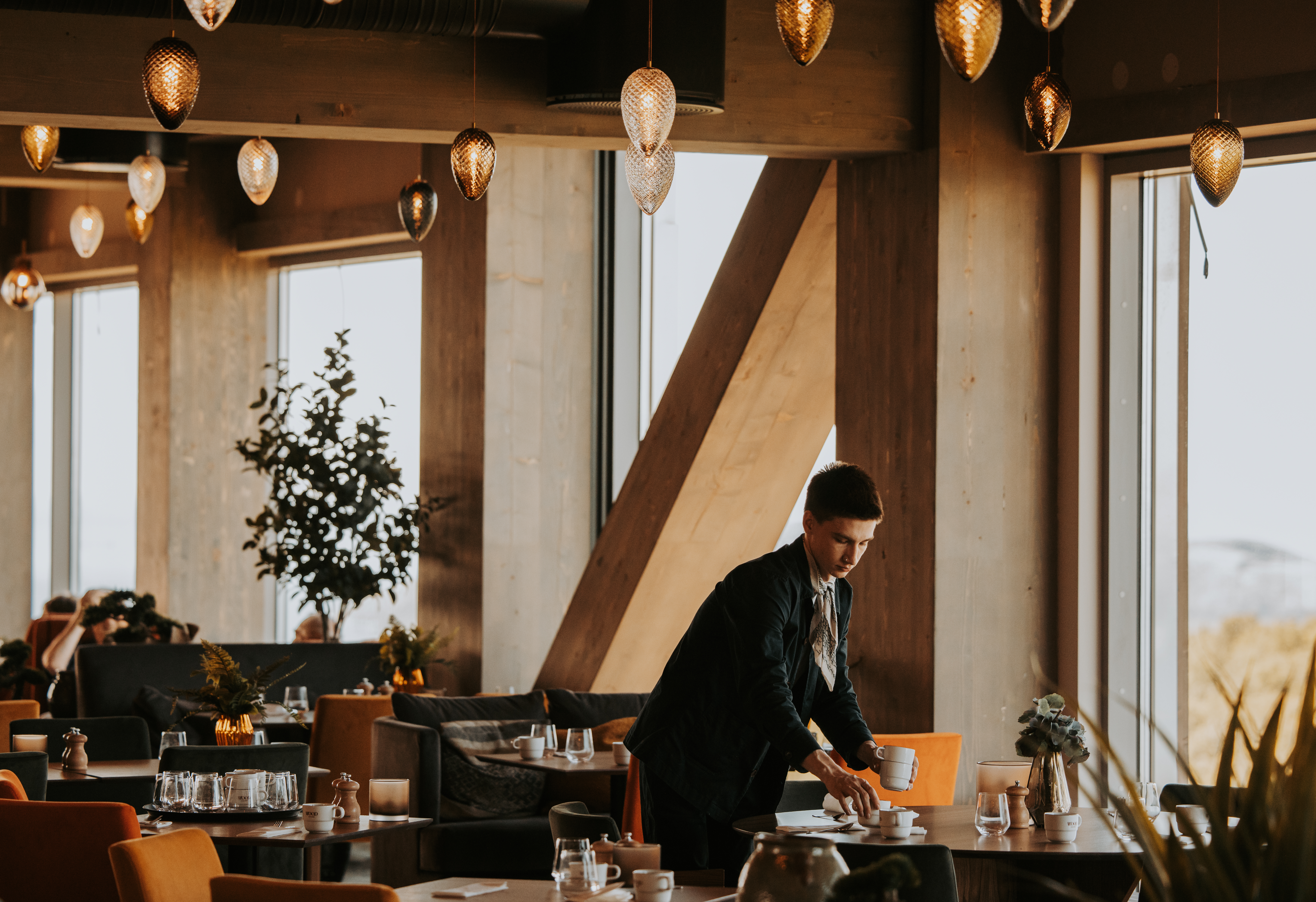 En person arrangerer et bord i en varmt oplyst, moderne restaurant med hængelamper og store vinduer. Bordene er dækket med glas og omgivet af hyggelige siddepladser.