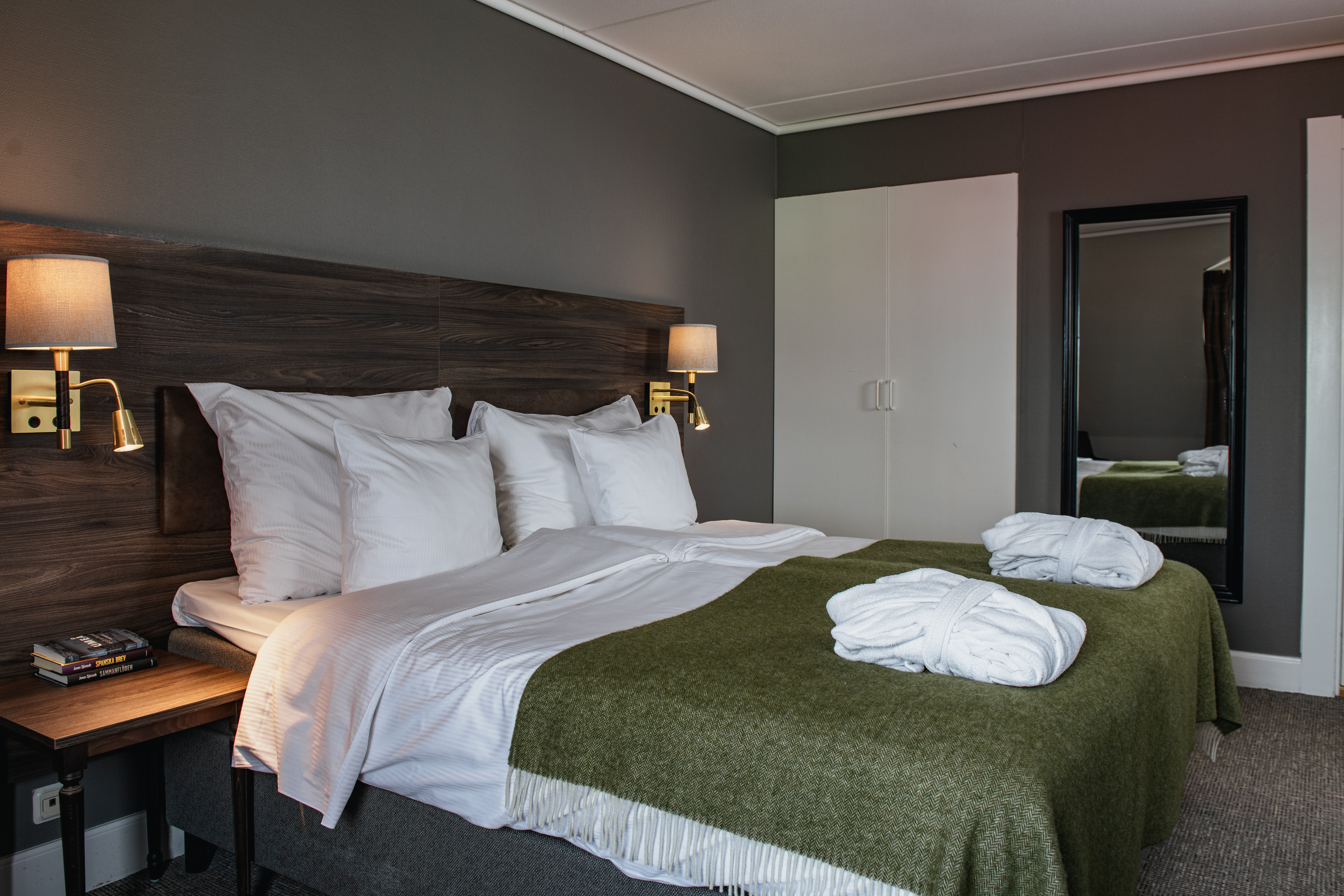 A neatly made bed with white pillows and a green blanket rests against a dark wooden headboard. Wall lamps illuminate the space, with a mirror and nightstand nearby.