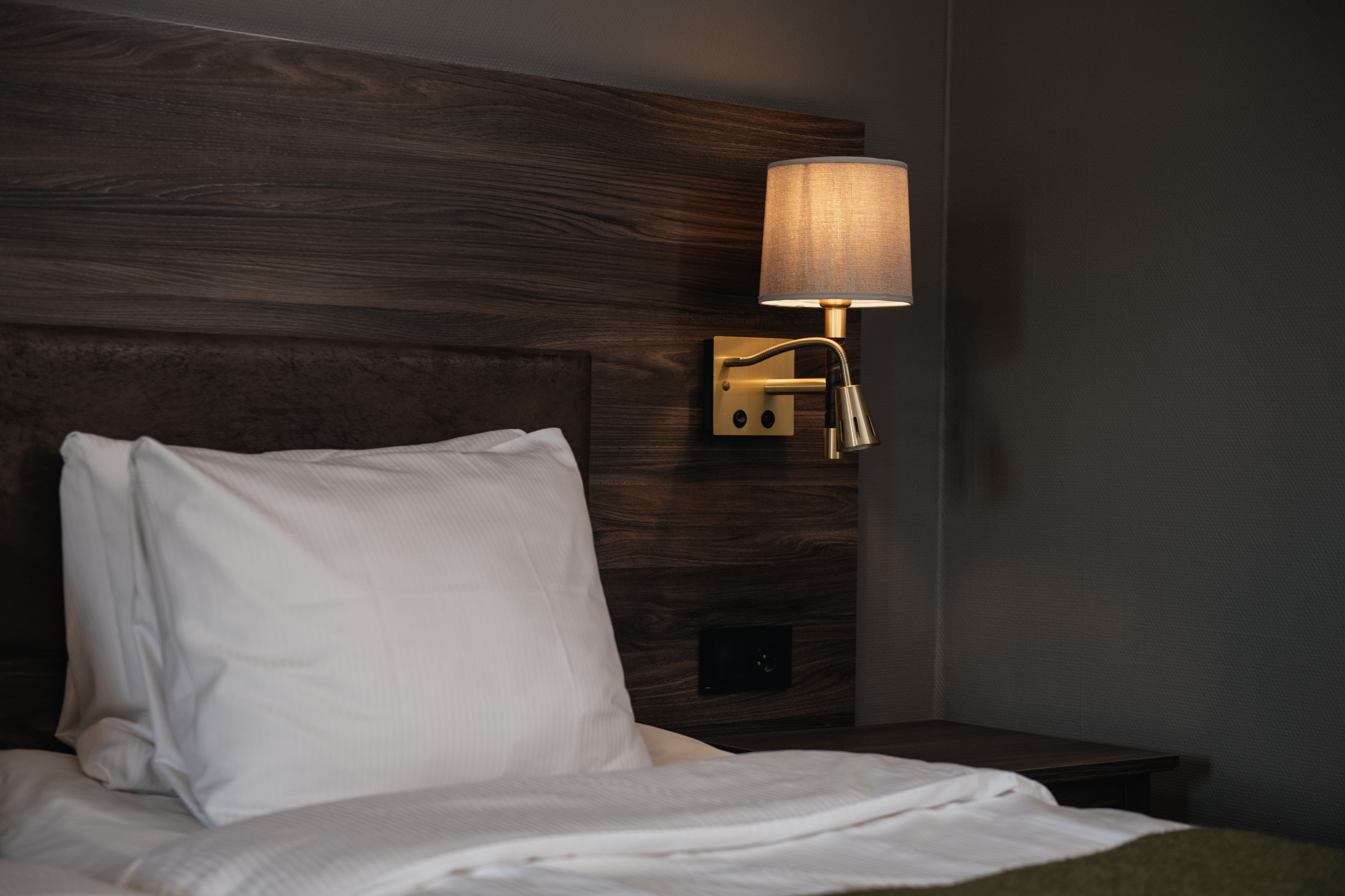Bed with white pillow and bedding, situated beneath a wooden headboard. A wall-mounted lamp with a beige shade illuminates the setting, creating a cozy bedroom atmosphere.