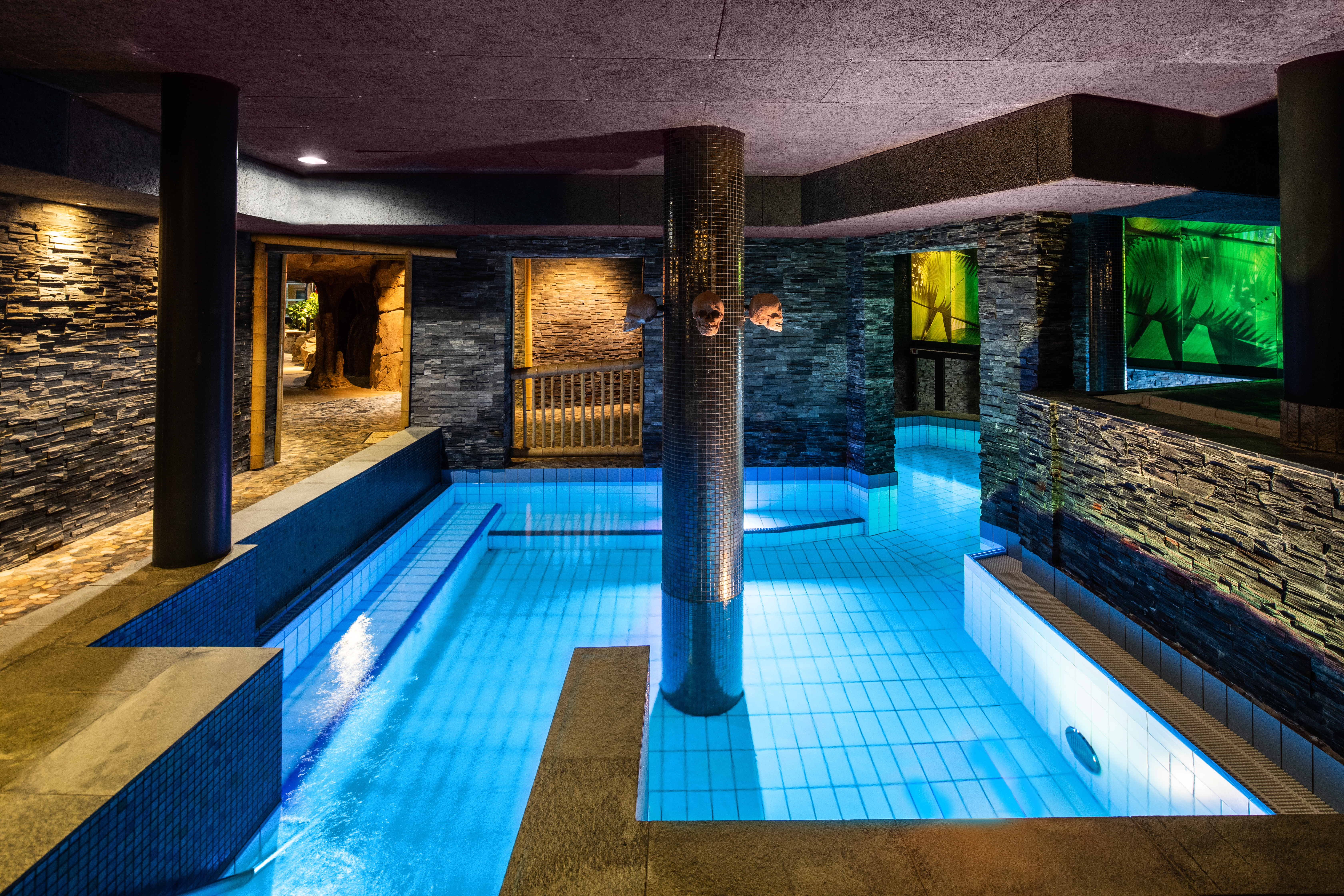 En indendørs pool med klart blåt vand omgiver en central søjle, omgivet af stenvægge og omgivende belysning. Døre og vinduer med udsigt til naturen forbedrer den rolige atmosfære.