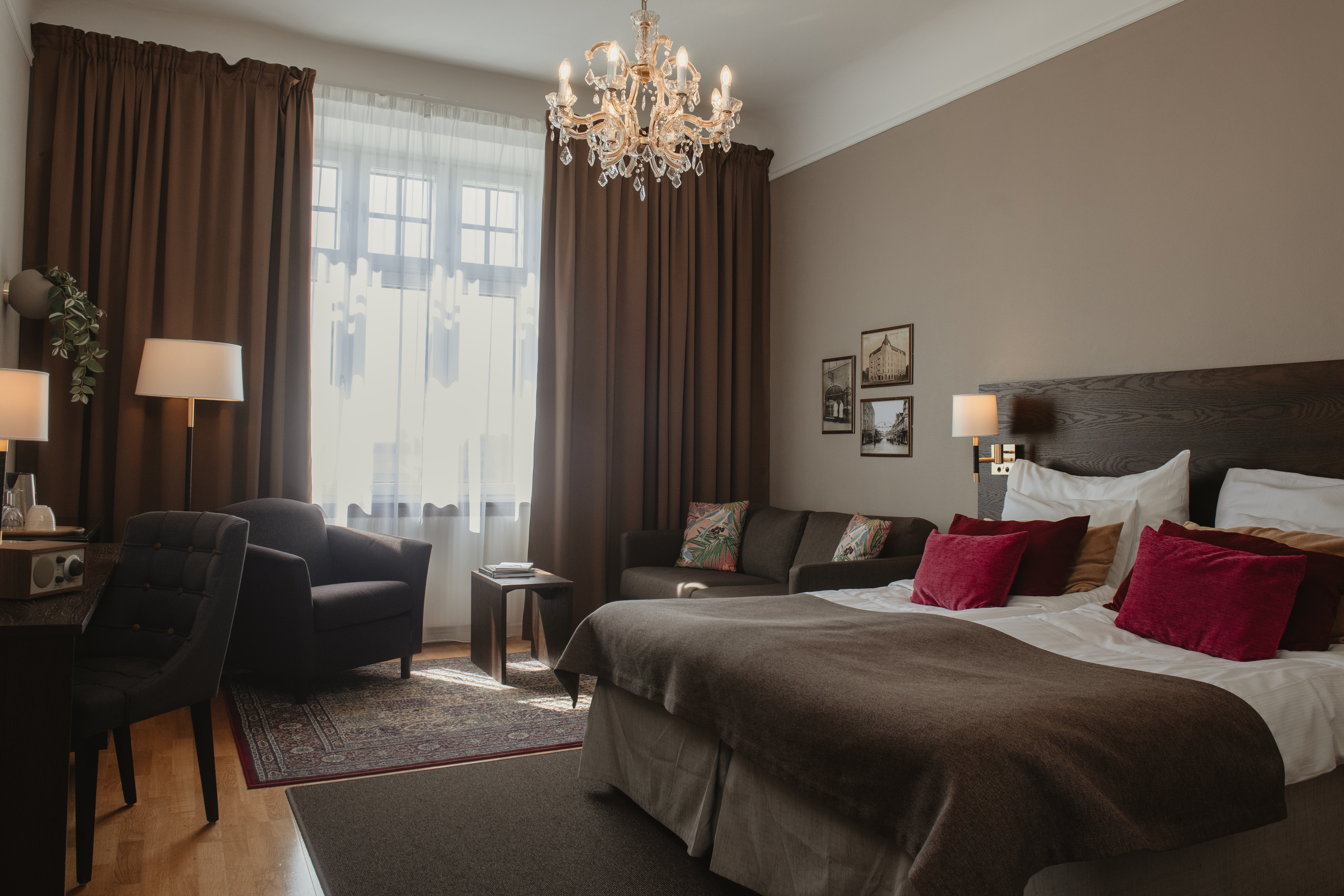Ett mysigt hotellrum har en stor säng med vinröda kuddar, en ljuskrona ovanför och en sittgrupp med stolar vid ett gardinbeklätt fönster; varm belysning förstärker den inbjudande atmosfären.