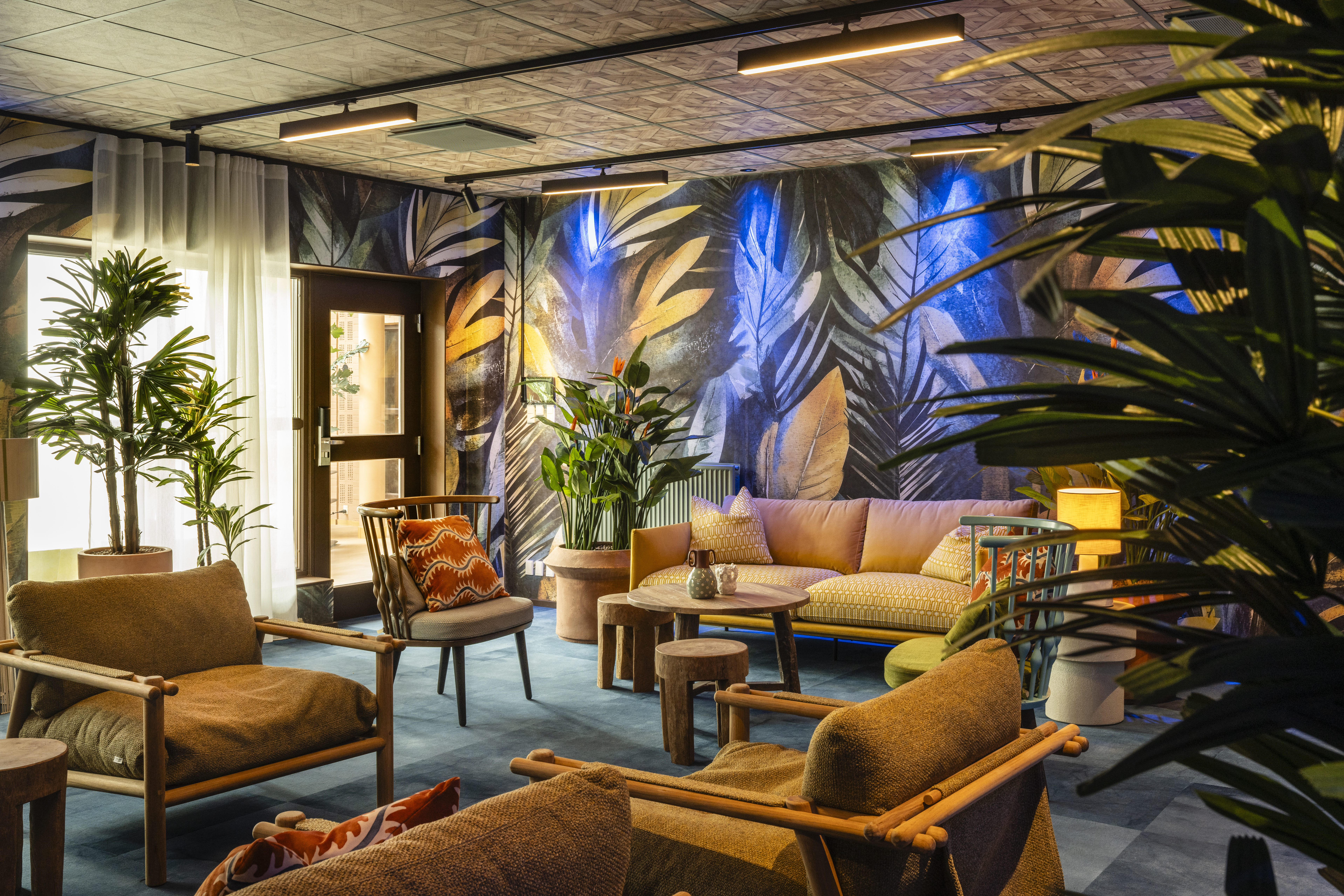 Komfortabel siddeområde med træstole og sofaer, med tropisk-inspireret tapet omgivet af frodige grønne planter, der skaber en hyggelig og livlig atmosfære.