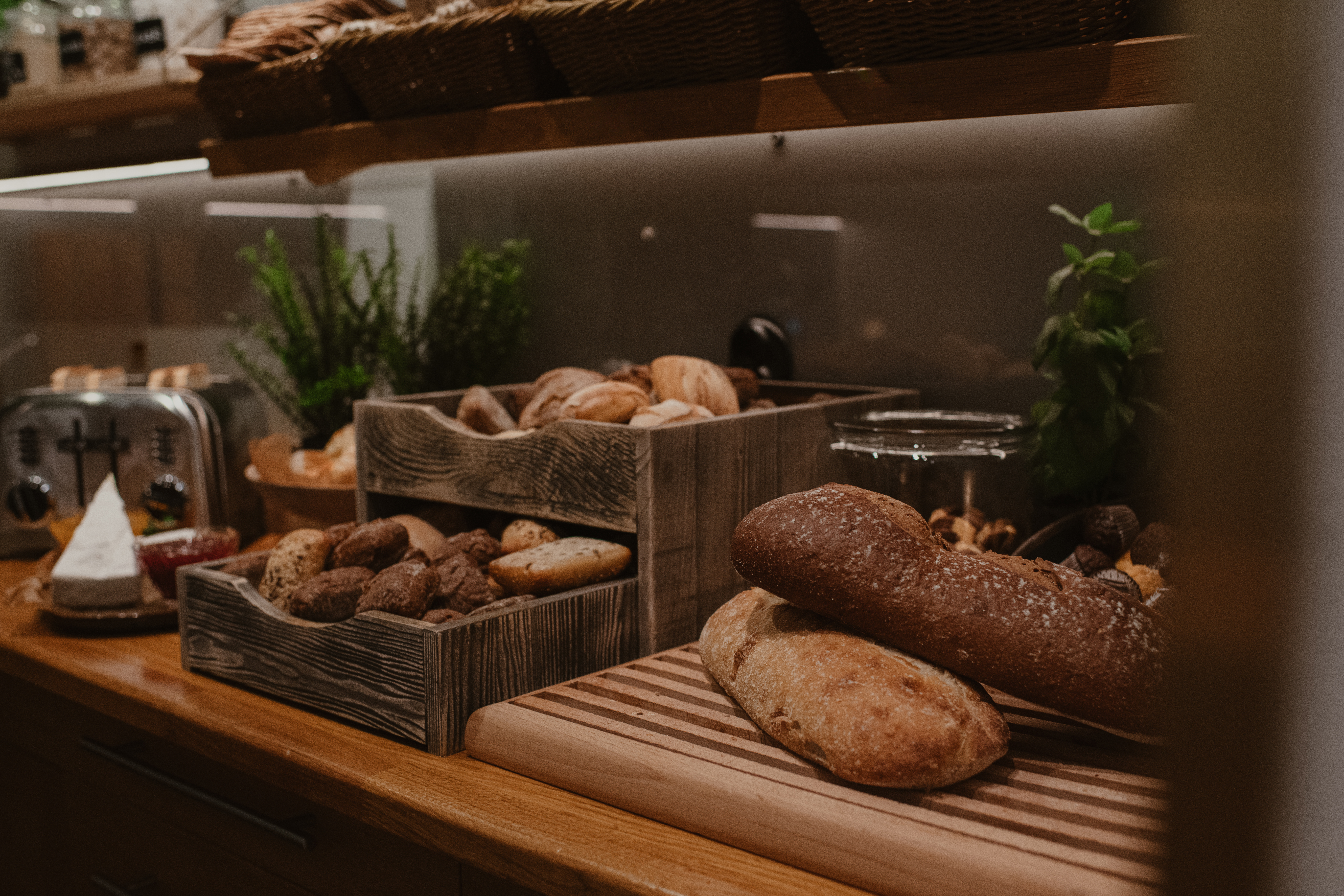 Brödlimpor och bullar på en träbänk med skärbrädor; krukväxter och en brödrost i bakgrunden skapar en varm, rustik köksmiljö.