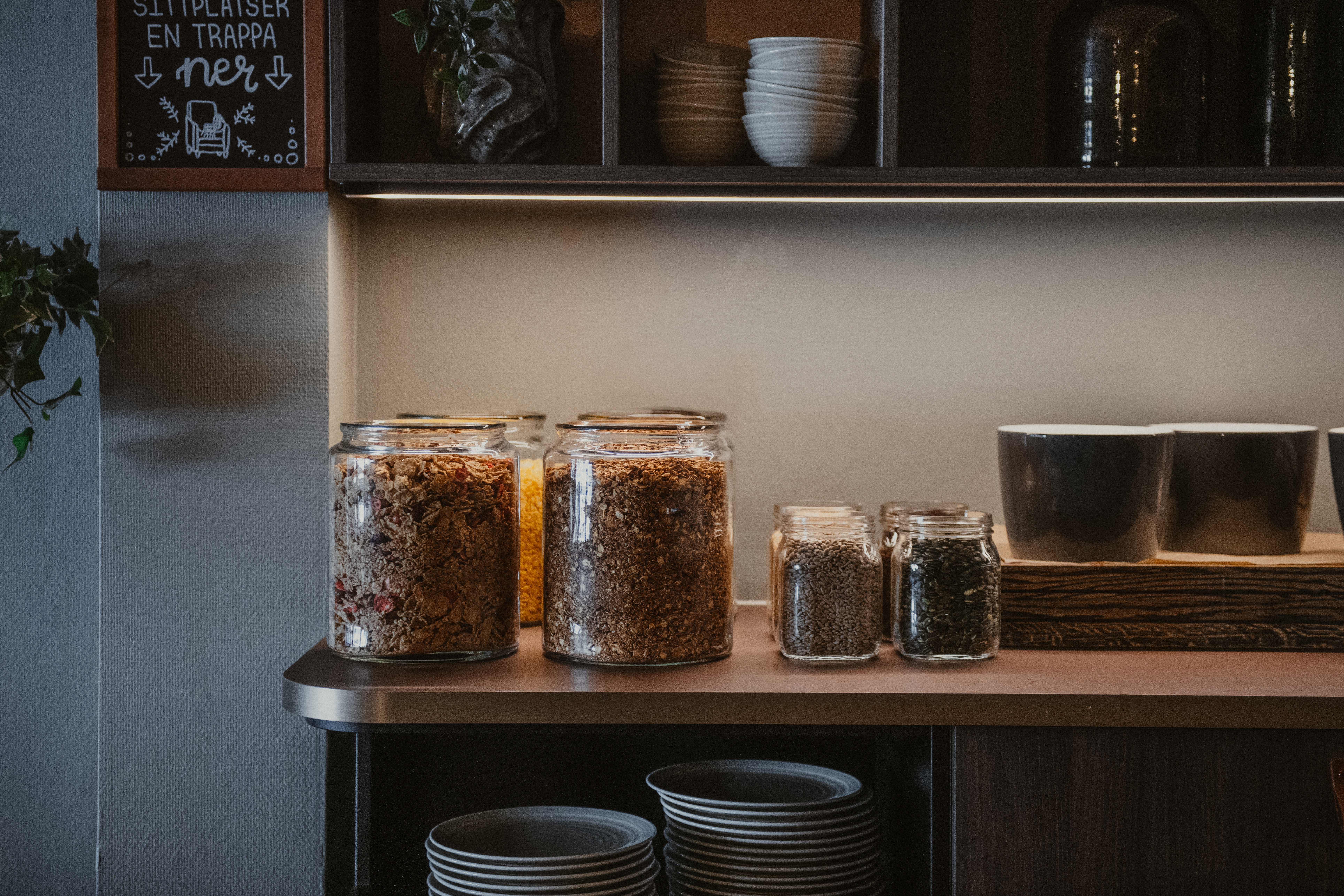 En självserveringsfrukoststation med burkar med granola och frön, koppar och tallrikar. En skylt lyder SITTPLATSER EN TRAPPA ner.
