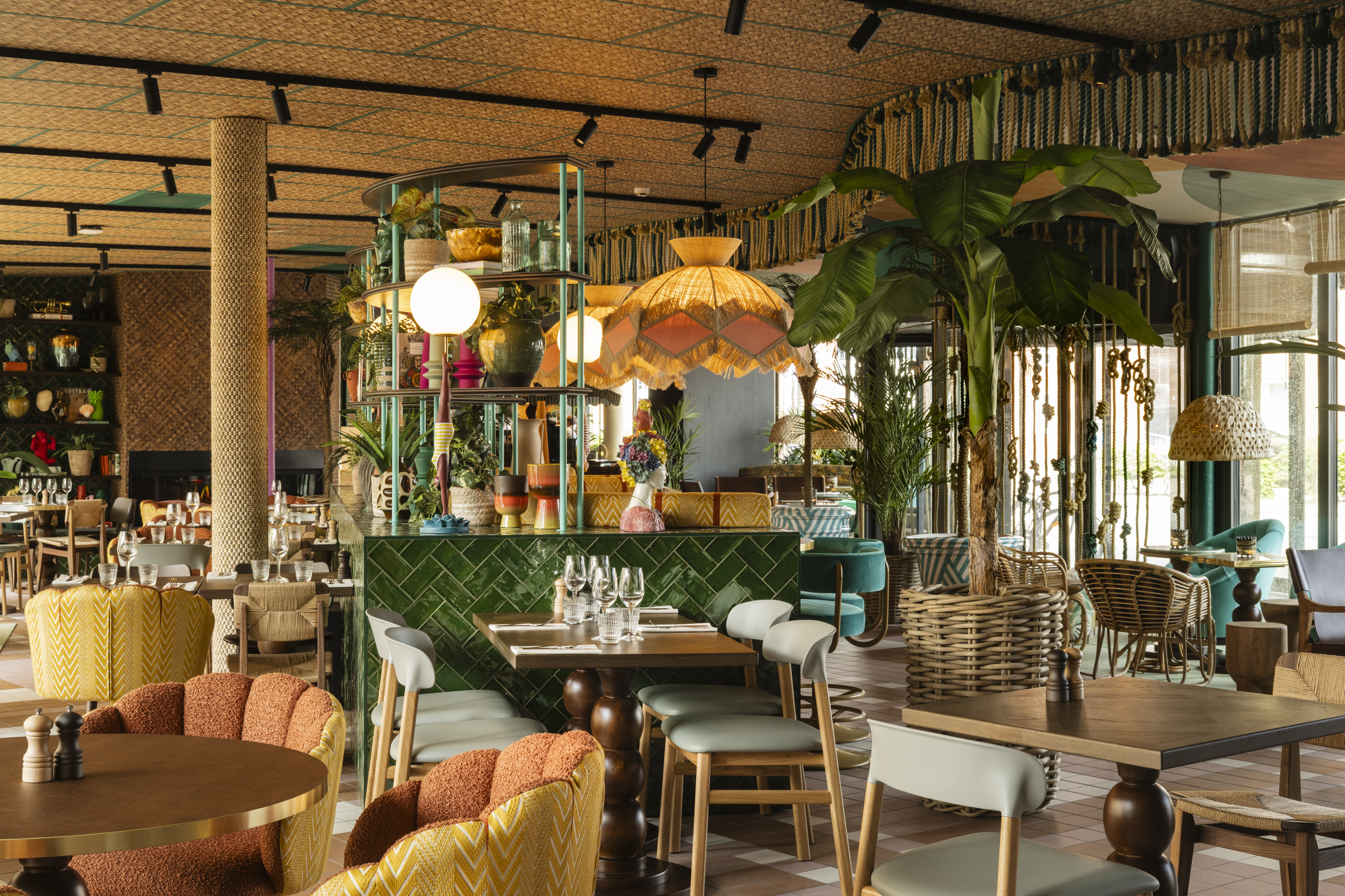 En restaurantindretning har farverig, eklektisk interiør med frodige planter, træmøbler og blød belysning. Bordene er sat med glas og bestik, hvilket skaber en indbydende spisestemning.