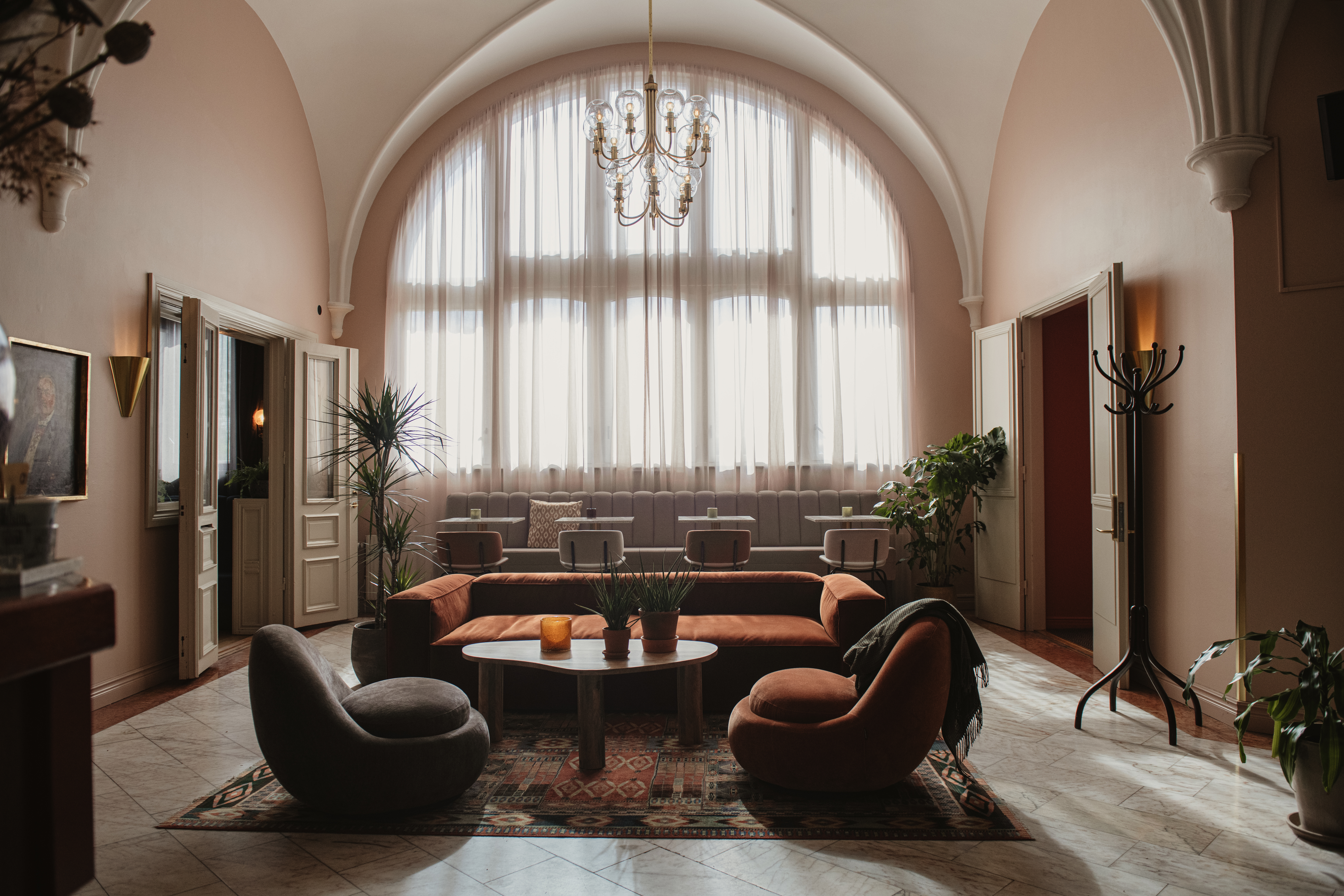 En hyggelig lounge har et rundt sofabord omgivet af moderne stole og en sofa. Sollys strømmer ind gennem store buede vinduer og belyser planter i et elegant rum.