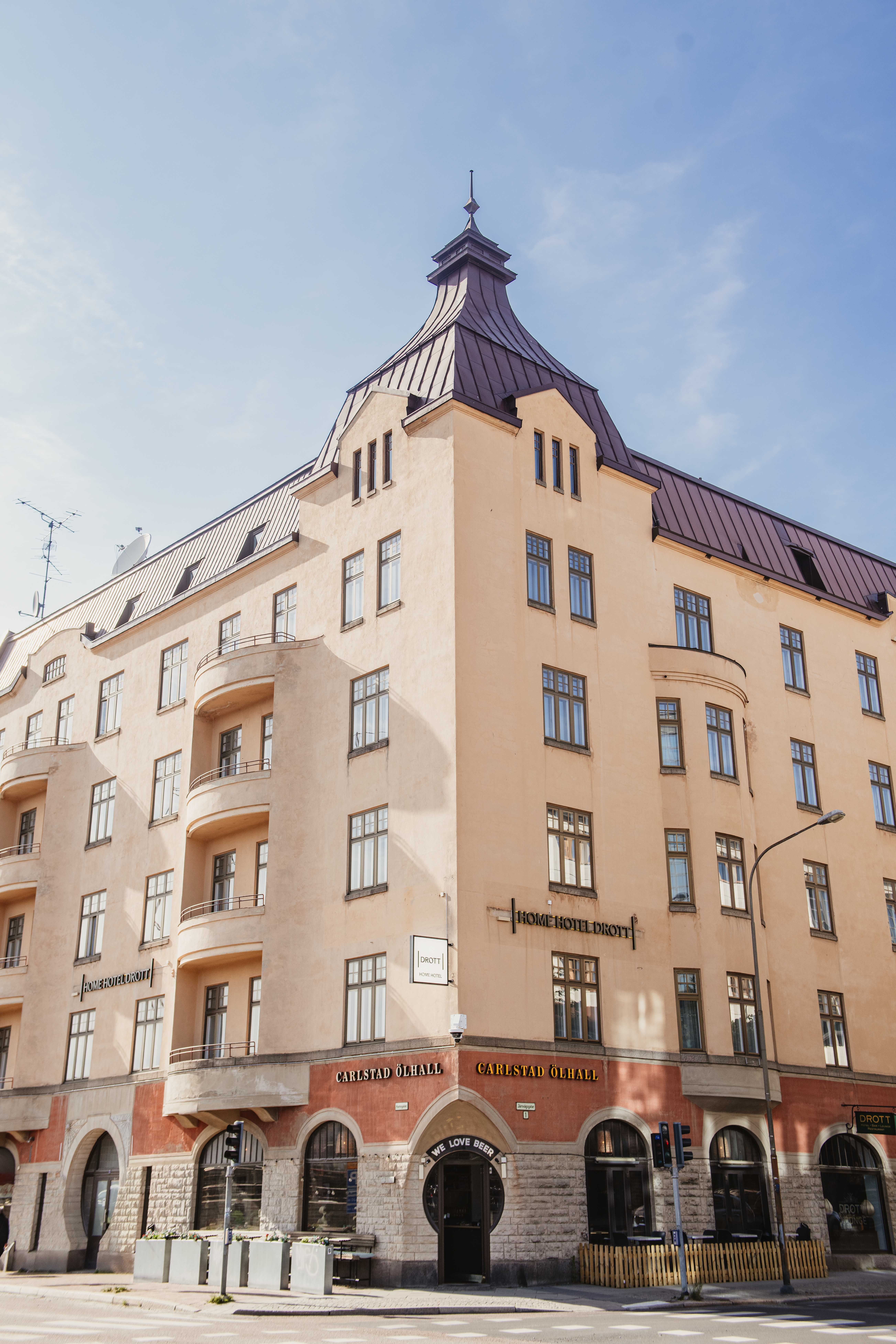 En beige flerbostadsbyggnad med mörkt tak står vid ett gathörn. Skyltar lyder "HOME HOTEL DROTT" och "CARLSTAD ÖLHALL". Balkonger är framträdande på ena sidan.