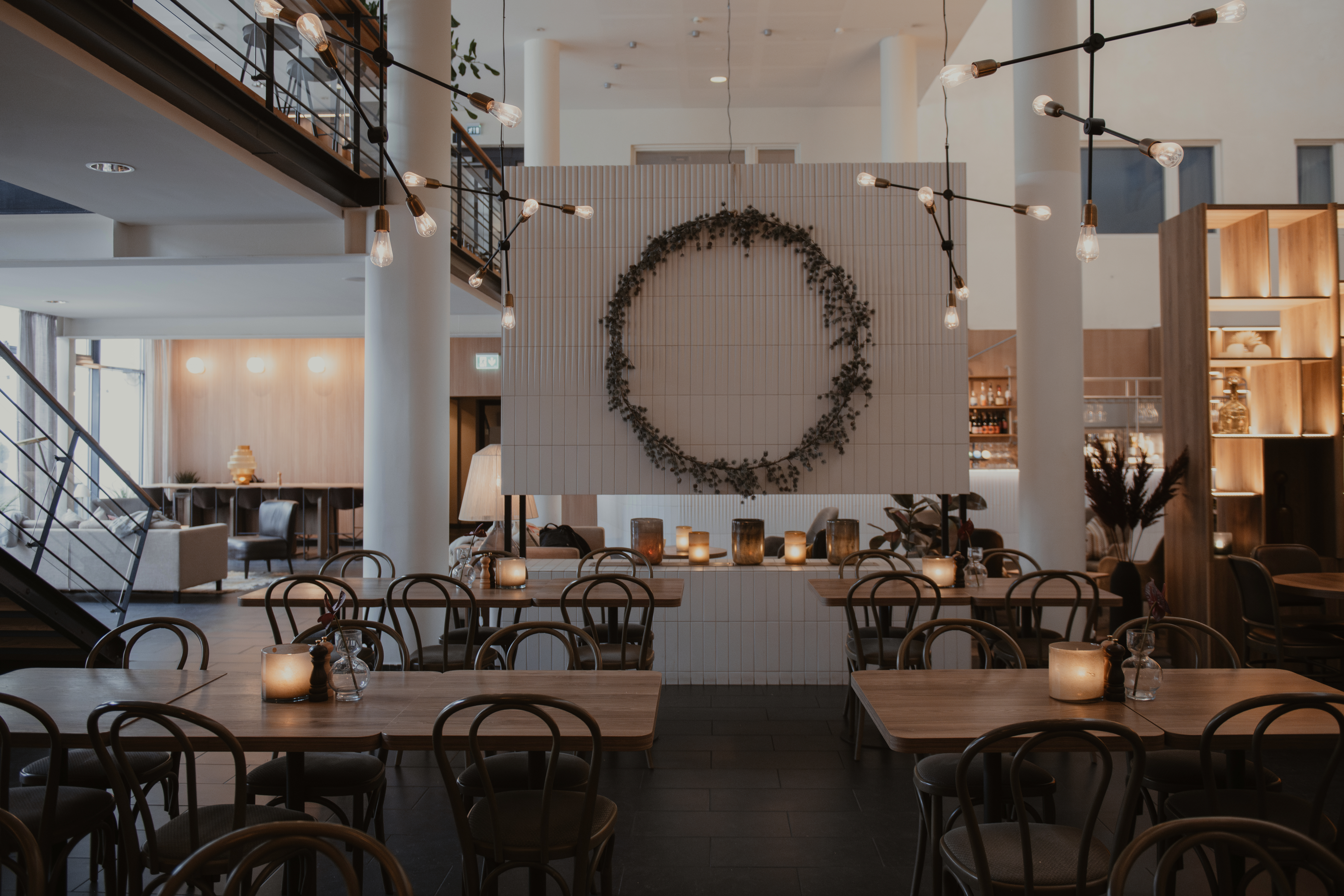 Matbord är ordnade med tända ljus i en modern, stilren restaurang. En stor dekorativ krans pryder en central vit vägg, omgiven av moderna sittplatser och belysningsarmaturer.