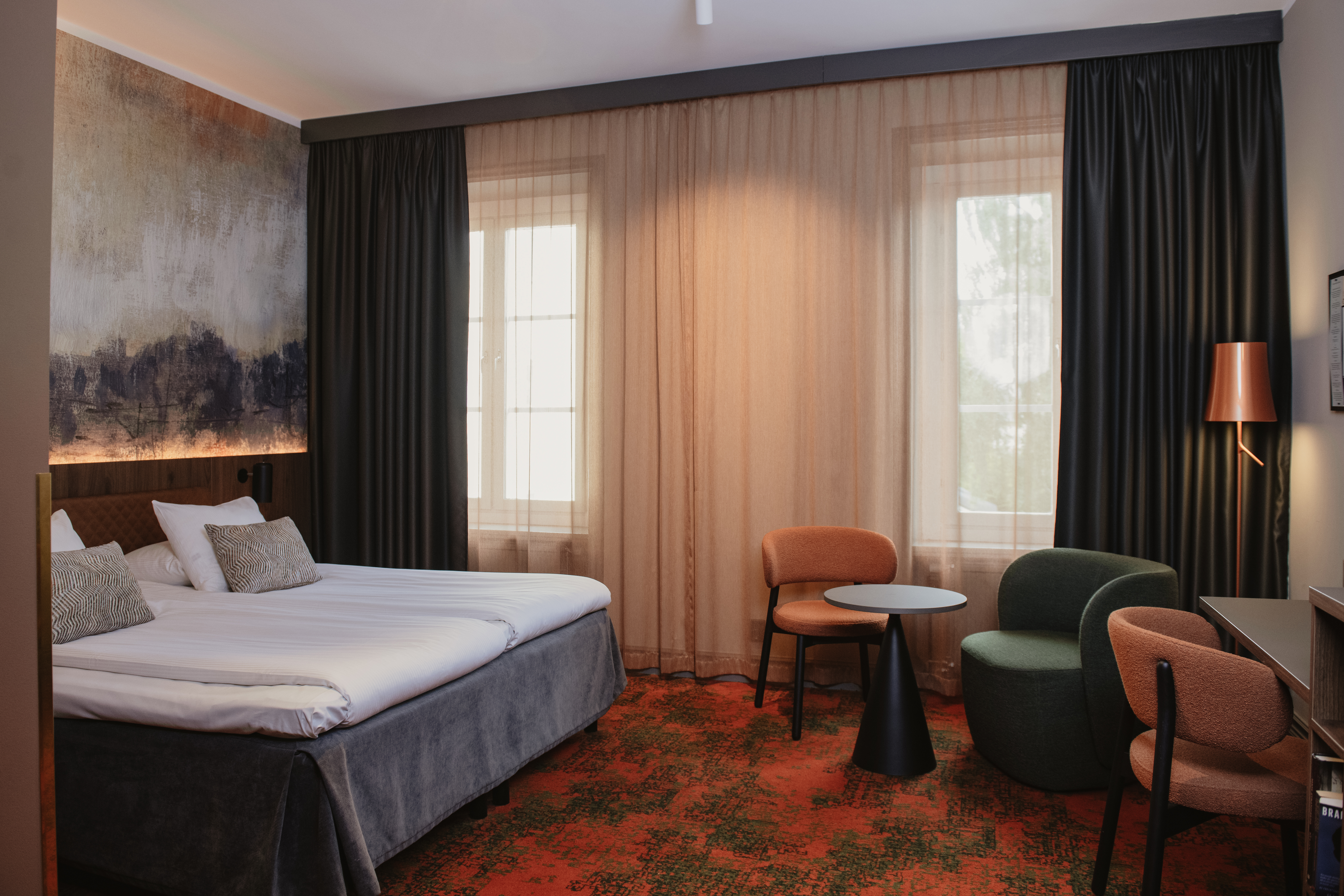 Et innbydende hotellrom med en komfortabel seng, kunstnerisk veggdekor og et koselig hjørne med stoler og et bord.