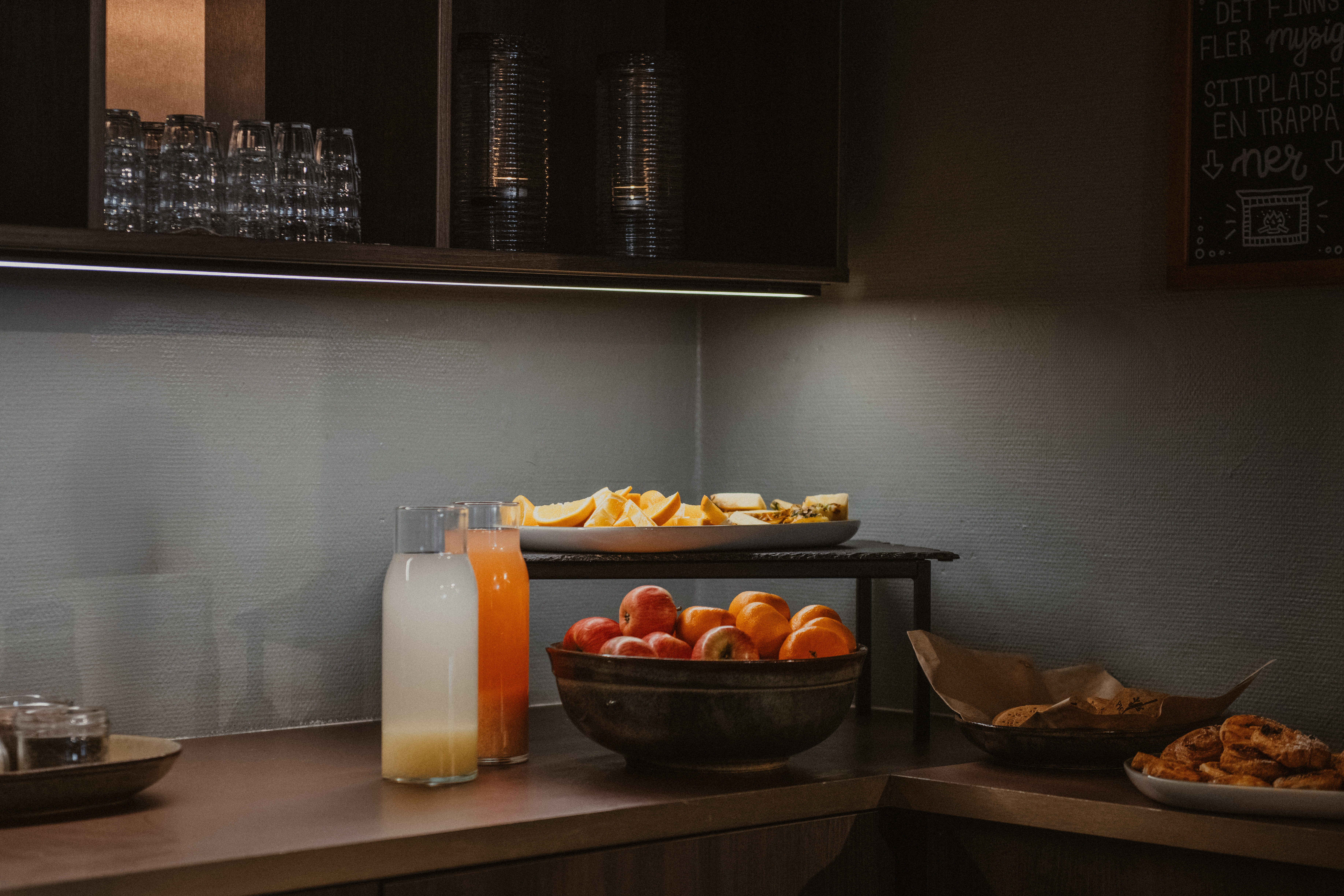 En självbetjäningsdisk med färsk juice, frukt och bakverk. Text på en griffeltavla lyder DET FINNS FLER mysig SITTPLATSER EN TRAPPA & ner.