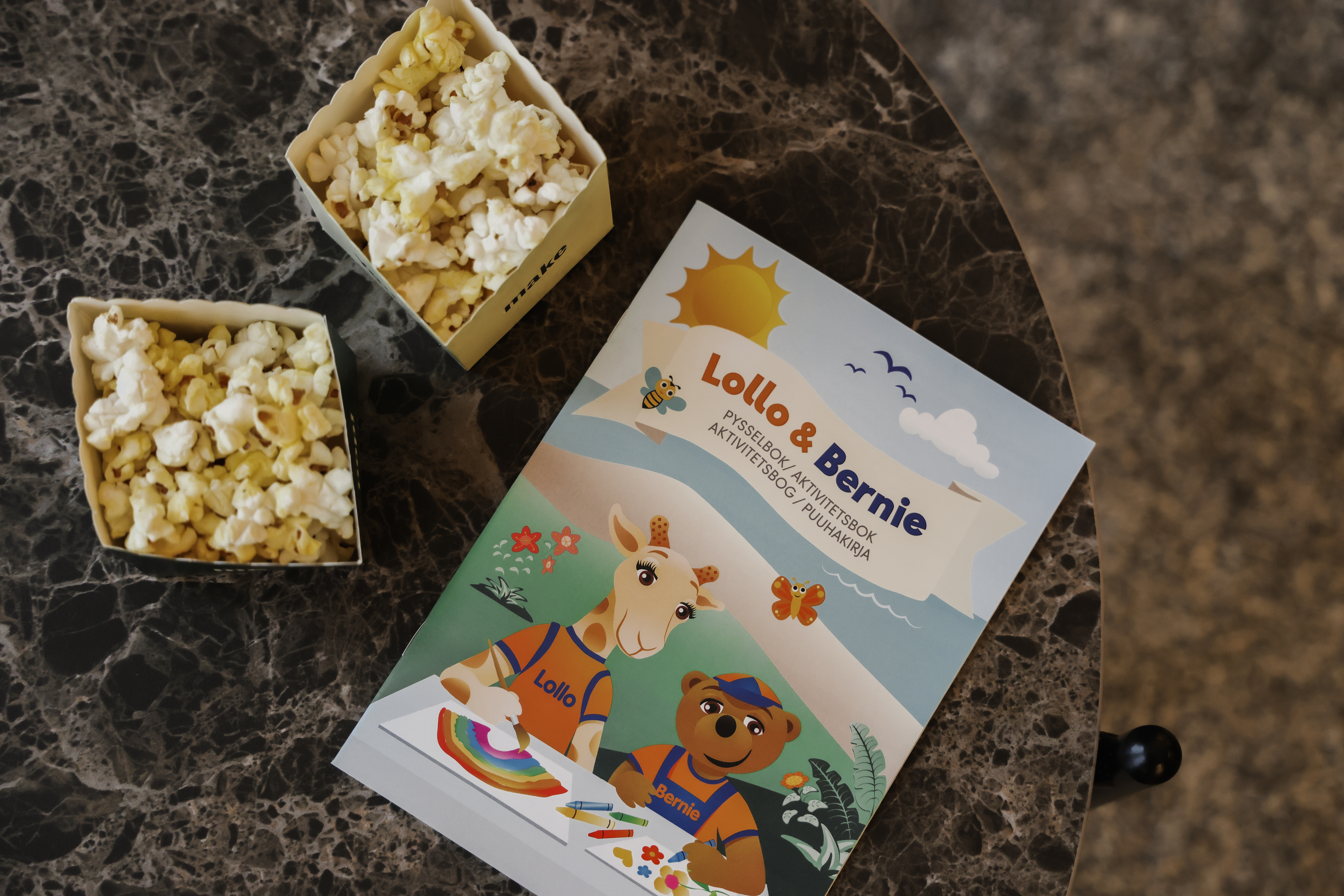 A children's activity booklet titled "Lollo & Bernie Activity Book" lies on a marble table beside two small open boxes of popcorn. The booklet features colorful illustrations of animals.