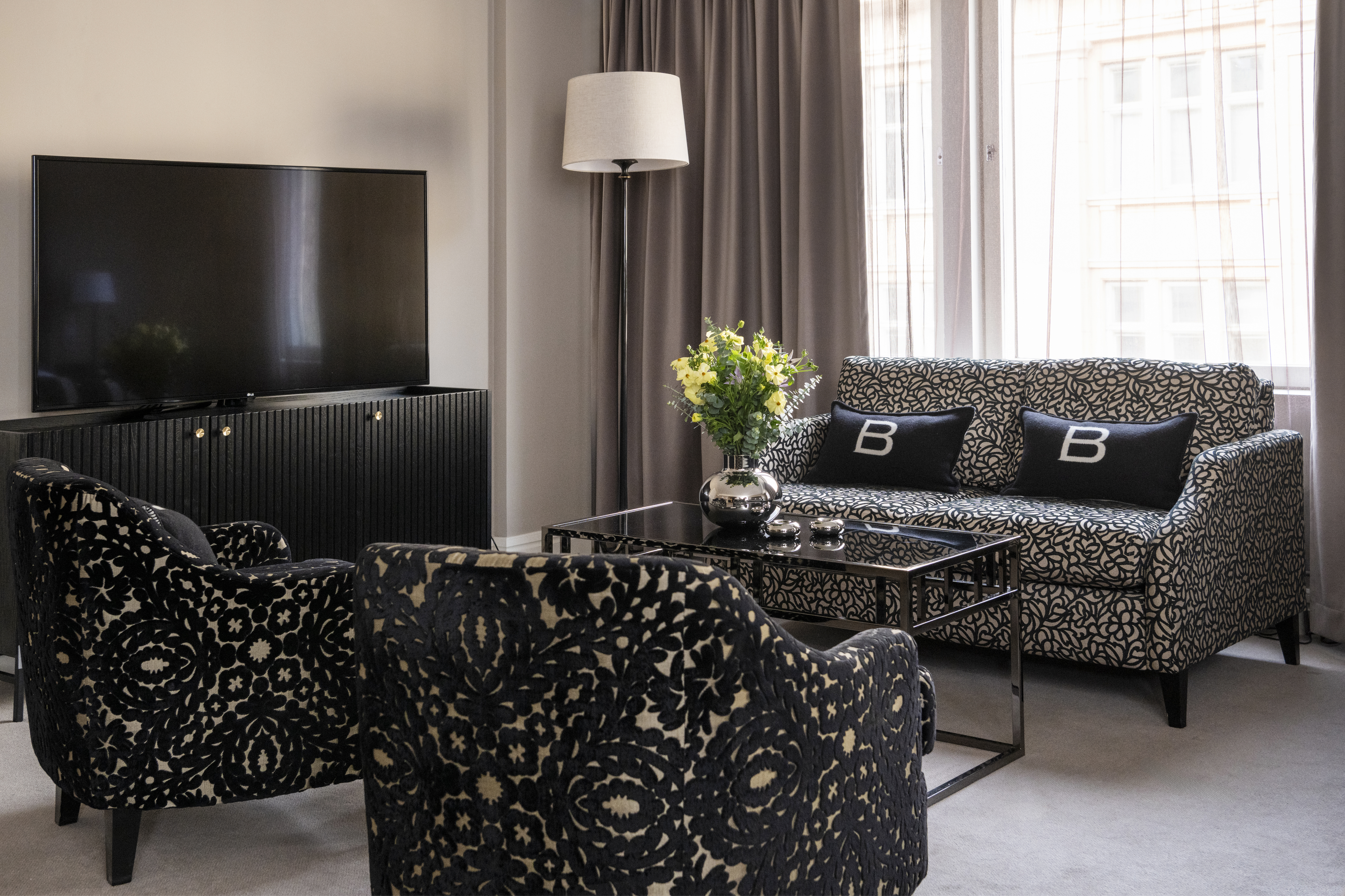 Television on a cabinet, facing patterned chairs and a sofa with "B" cushions. Flowers on glass coffee table; windows with curtains illuminate the room.