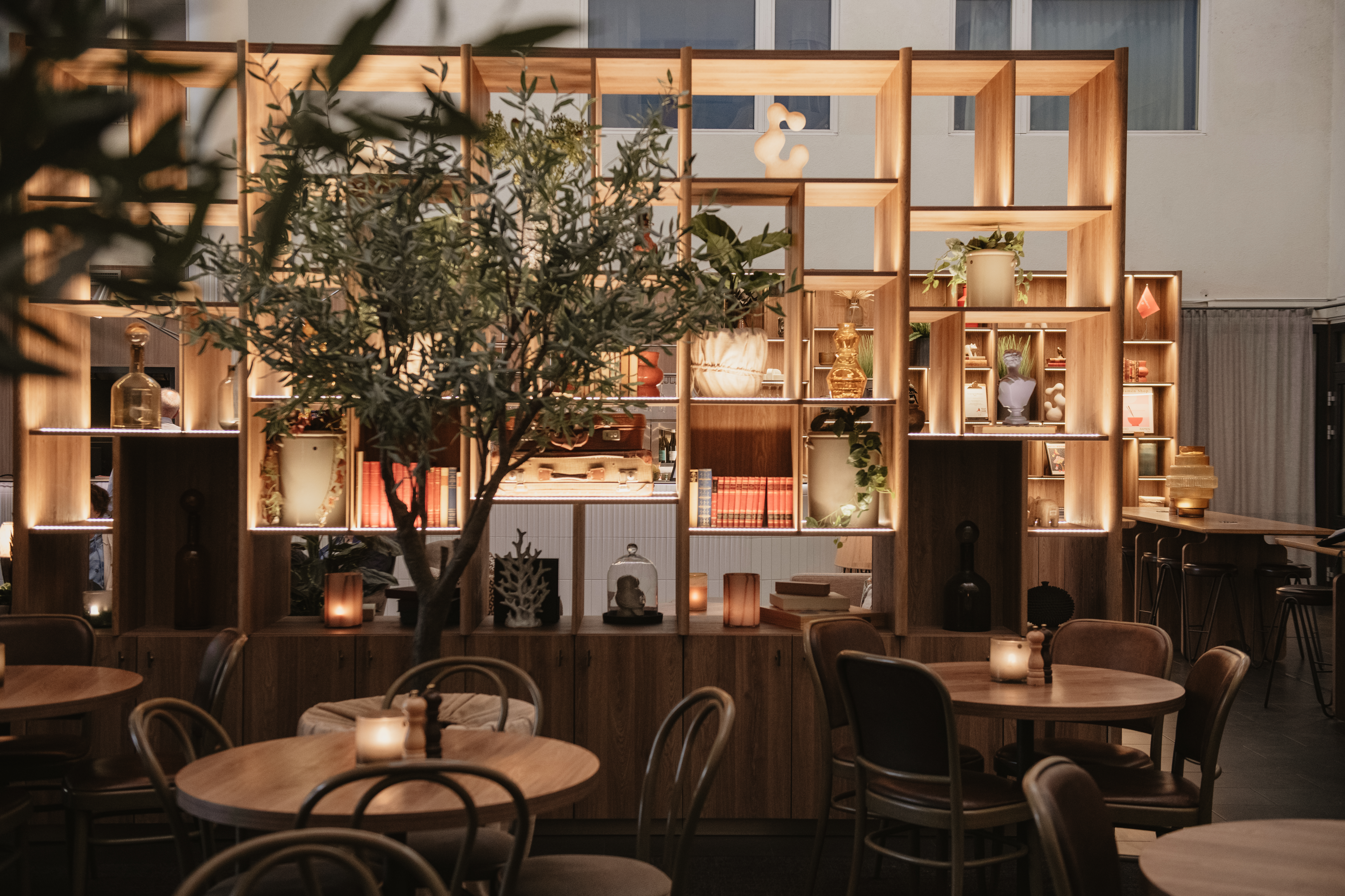 Bord med stearinlys er arrangerede foran en træhylde, der viser vaser og dekorationer, sat i en varm oplyst, hyggelig café eller restaurant, med et potte træ.