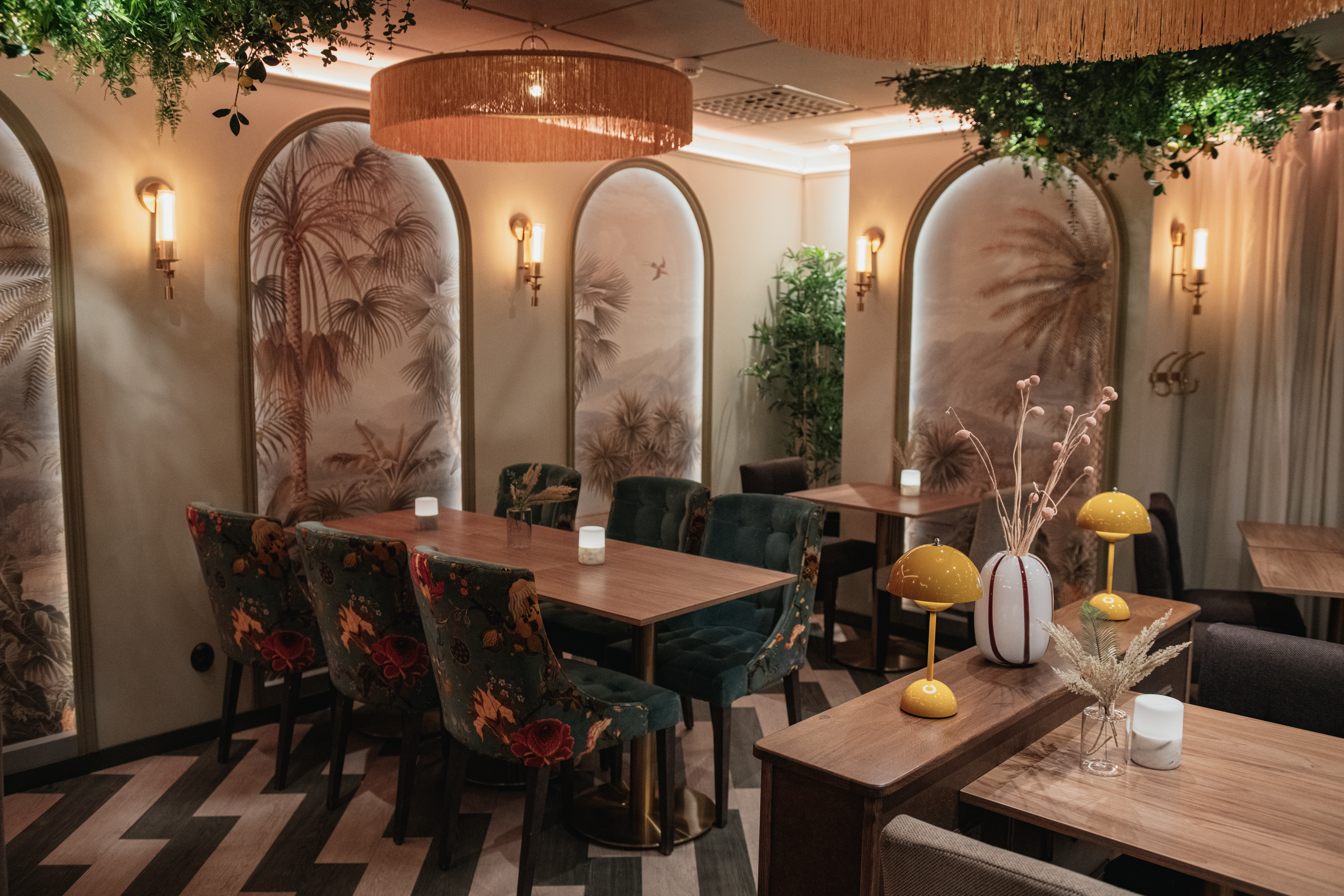 Matbord med blommiga stolar, i en varm, mysig restaurang. Tropiska väggkonstverk och hängande grönska skapar en inbjudande atmosfär, med mjuk belysning från vägglampor och taklampor.