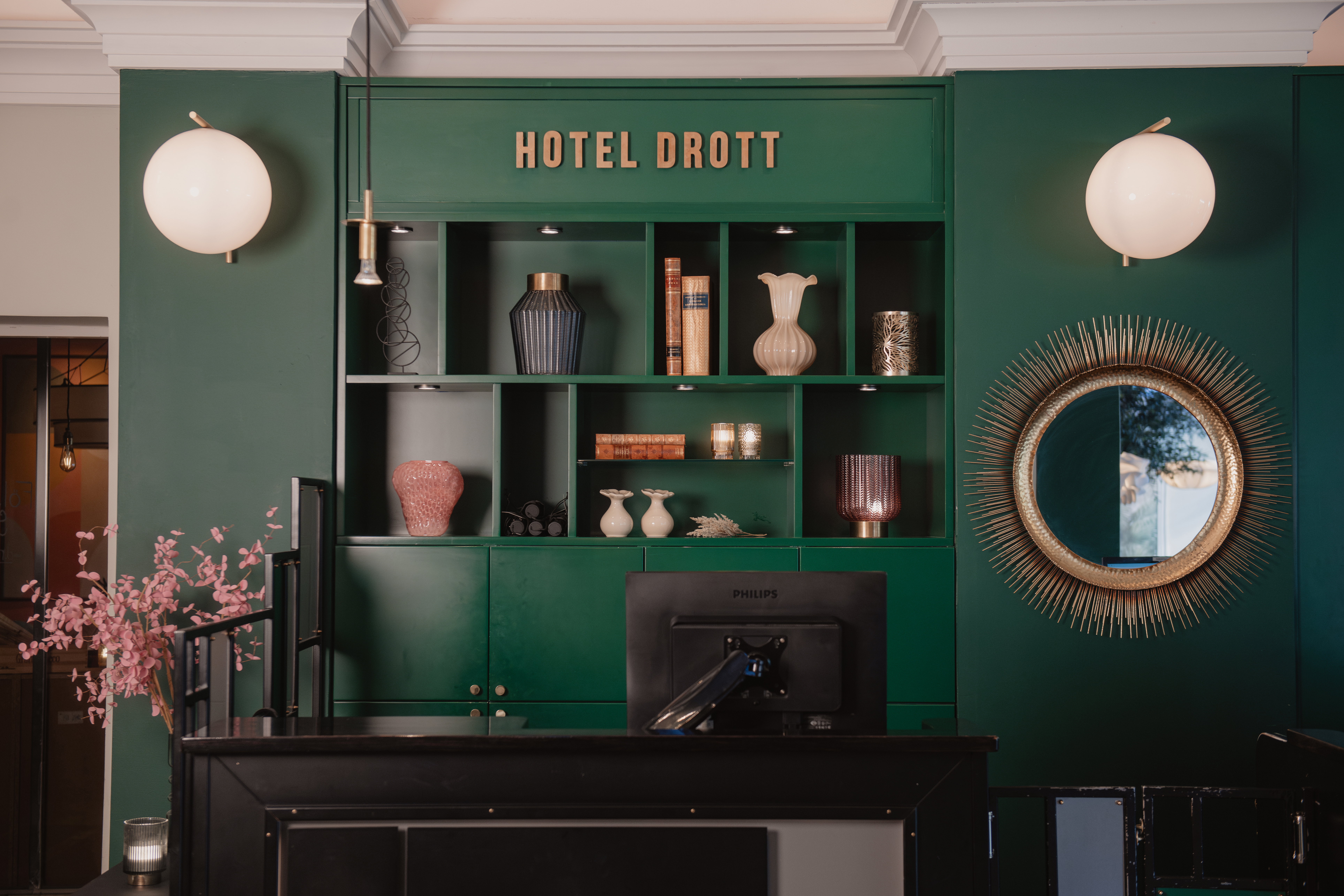 Reception med rosa blommor framför en grön hylla med vaser och böcker. "HOTEL DROTT" står skrivet ovanför. Runda speglar och lampor pryder väggarna.