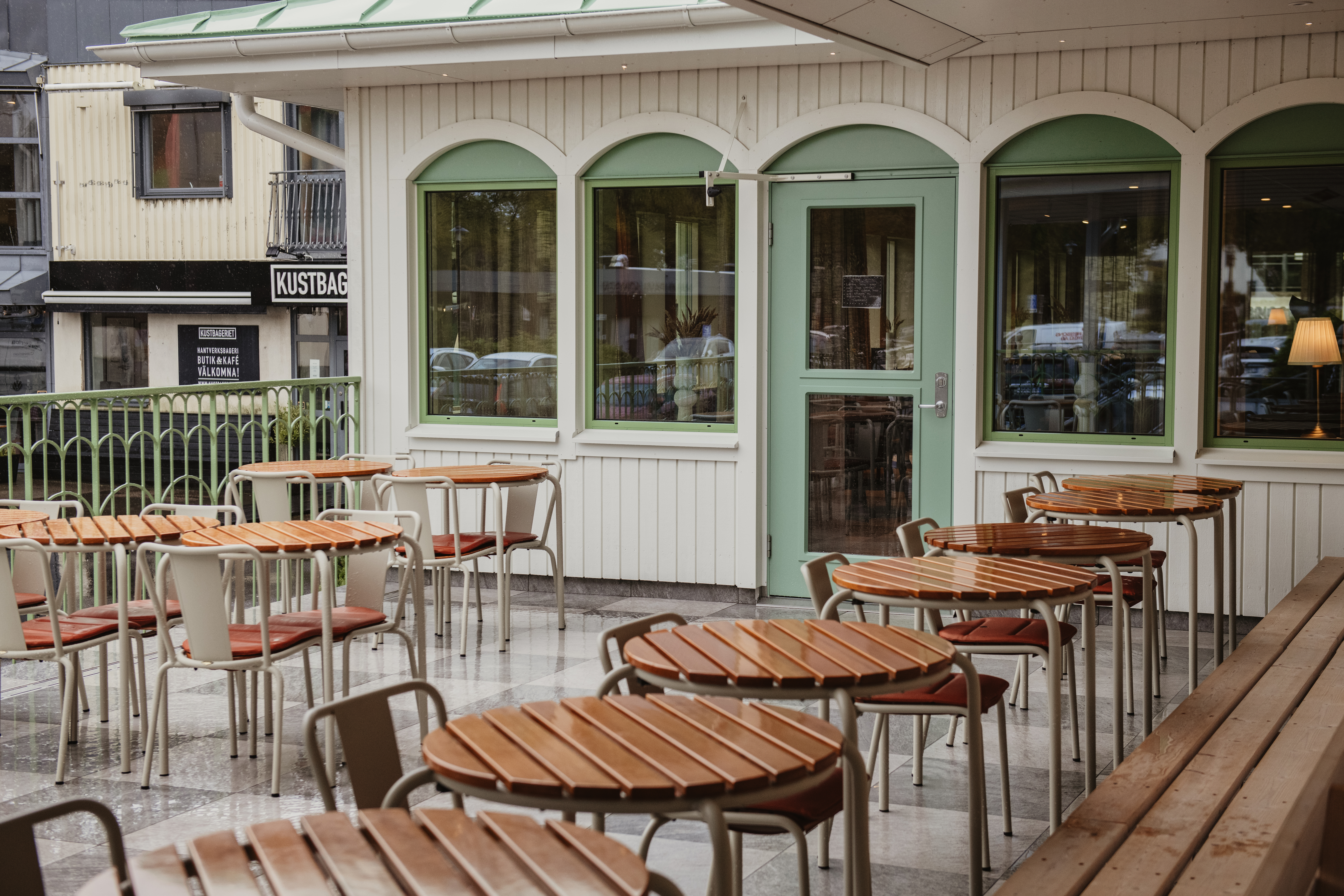 En udendørs caféterrasse med mange borde og stole, ved siden af en bygning med grønrammede vinduer. Et skilt læser OCH KAFE VALKOMNA.