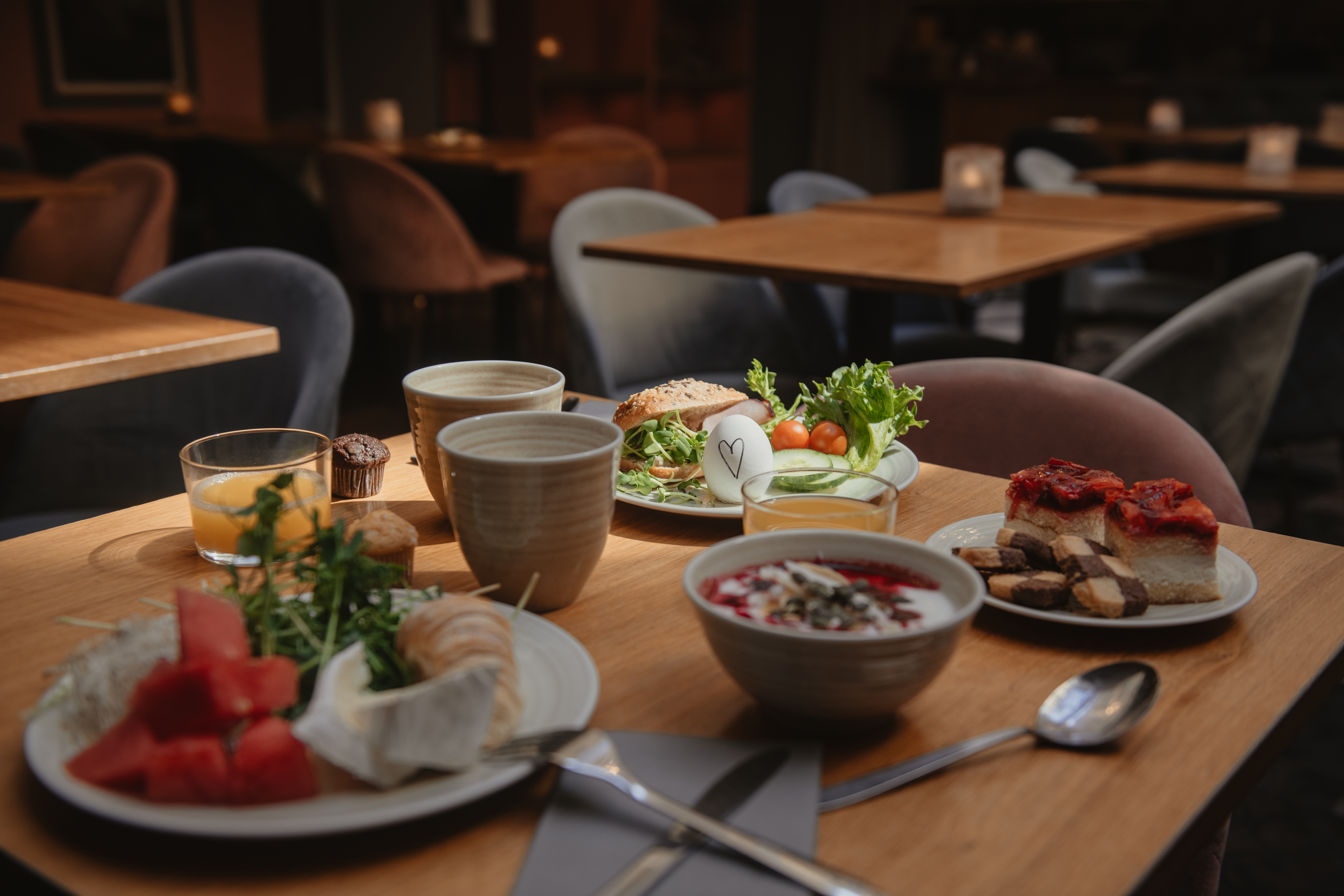 Ett träbord är dukat med frukostmat – frukt, yoghurt, smörgåsar och bakverk – omgiven av klädda stolar i ett varmt upplyst matområde.