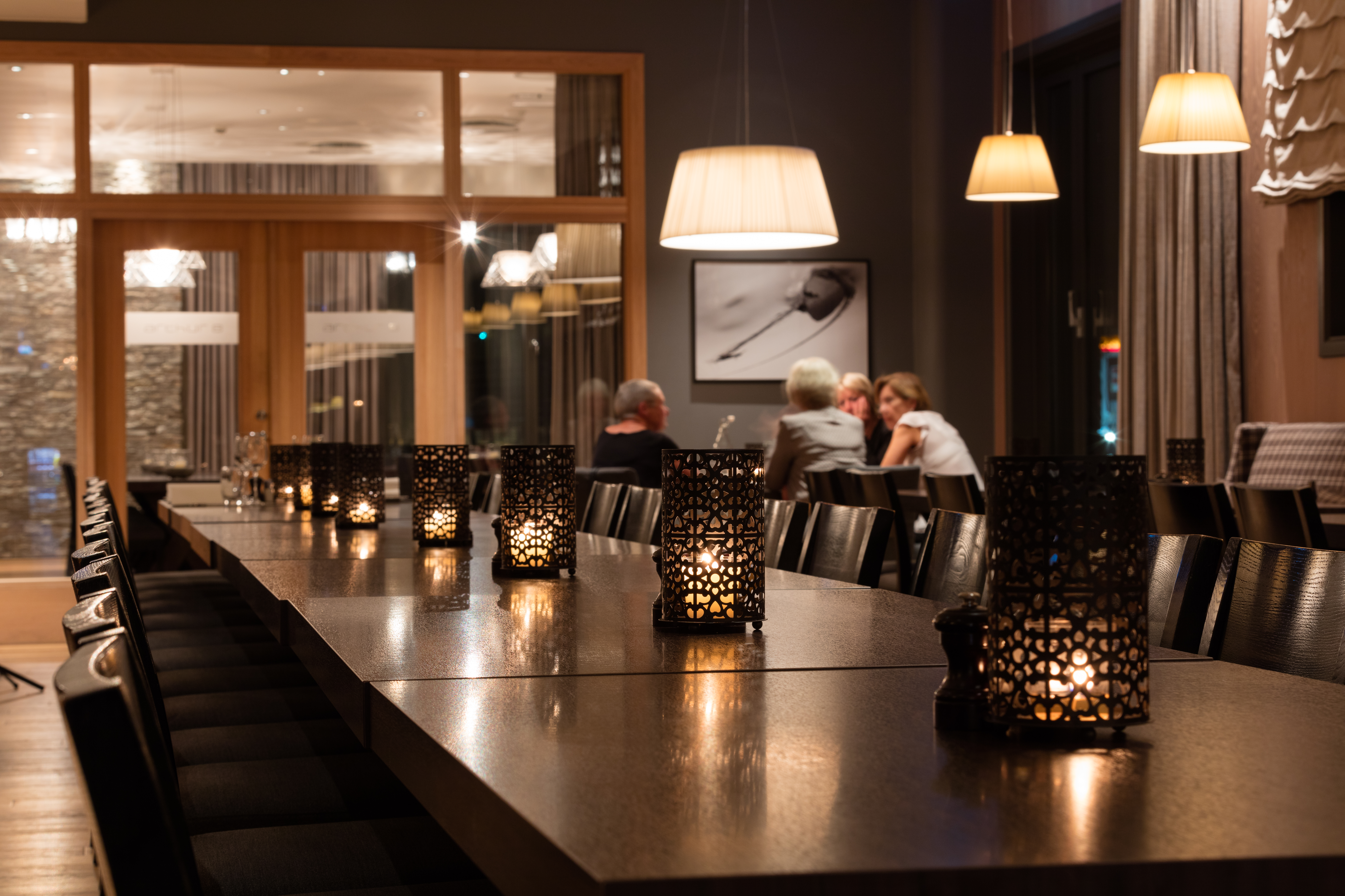 Stearinlys lyser opp et langt trebord, og skaper en varm atmosfære i en moderne restaurant. Folk samtaler i bakgrunnen under store pendellamper.