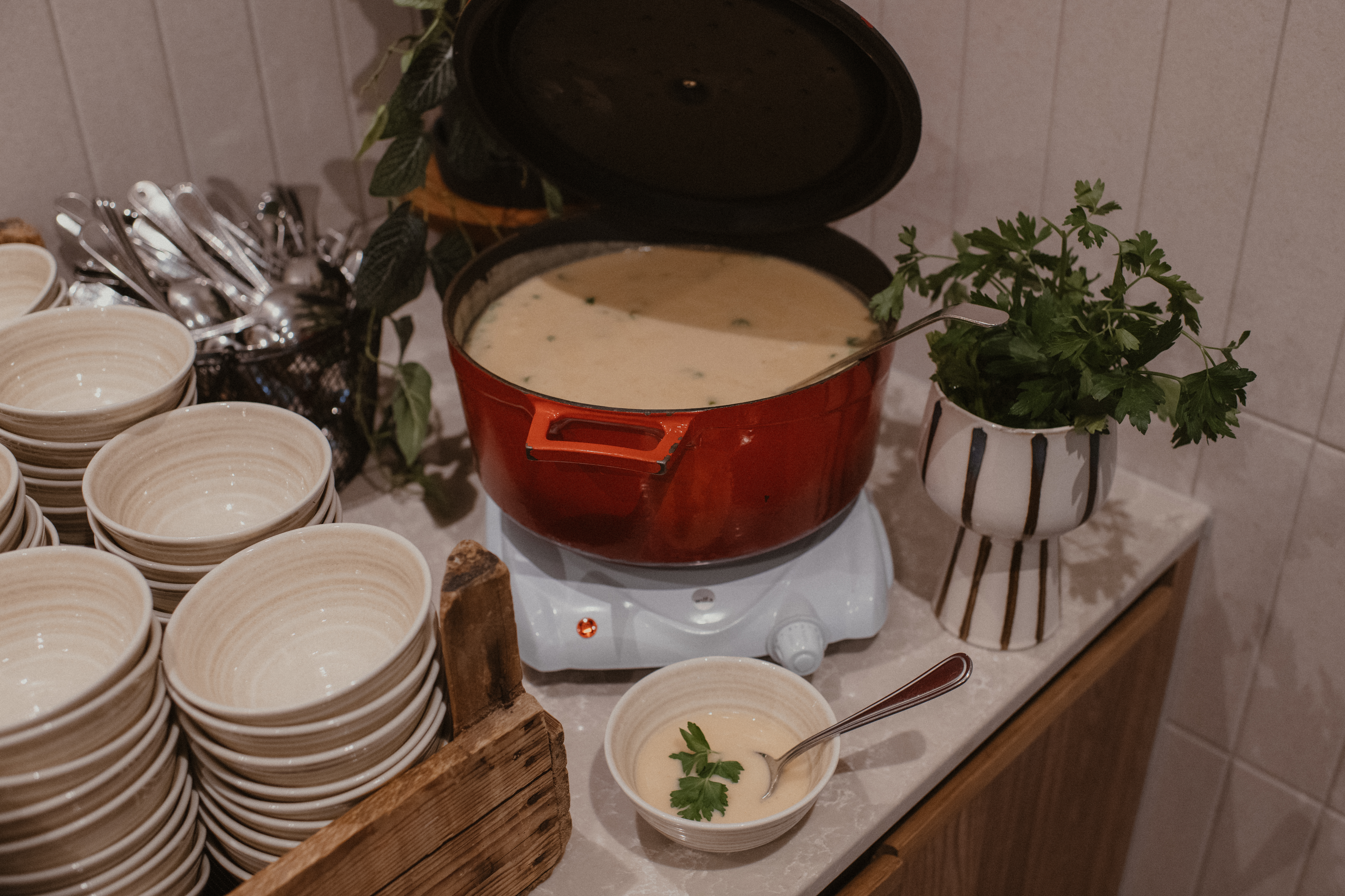 En röd gryta med krämig soppa värms på en platta, omgiven av staplade skålar och färsk persilja i en kruka på en disk.