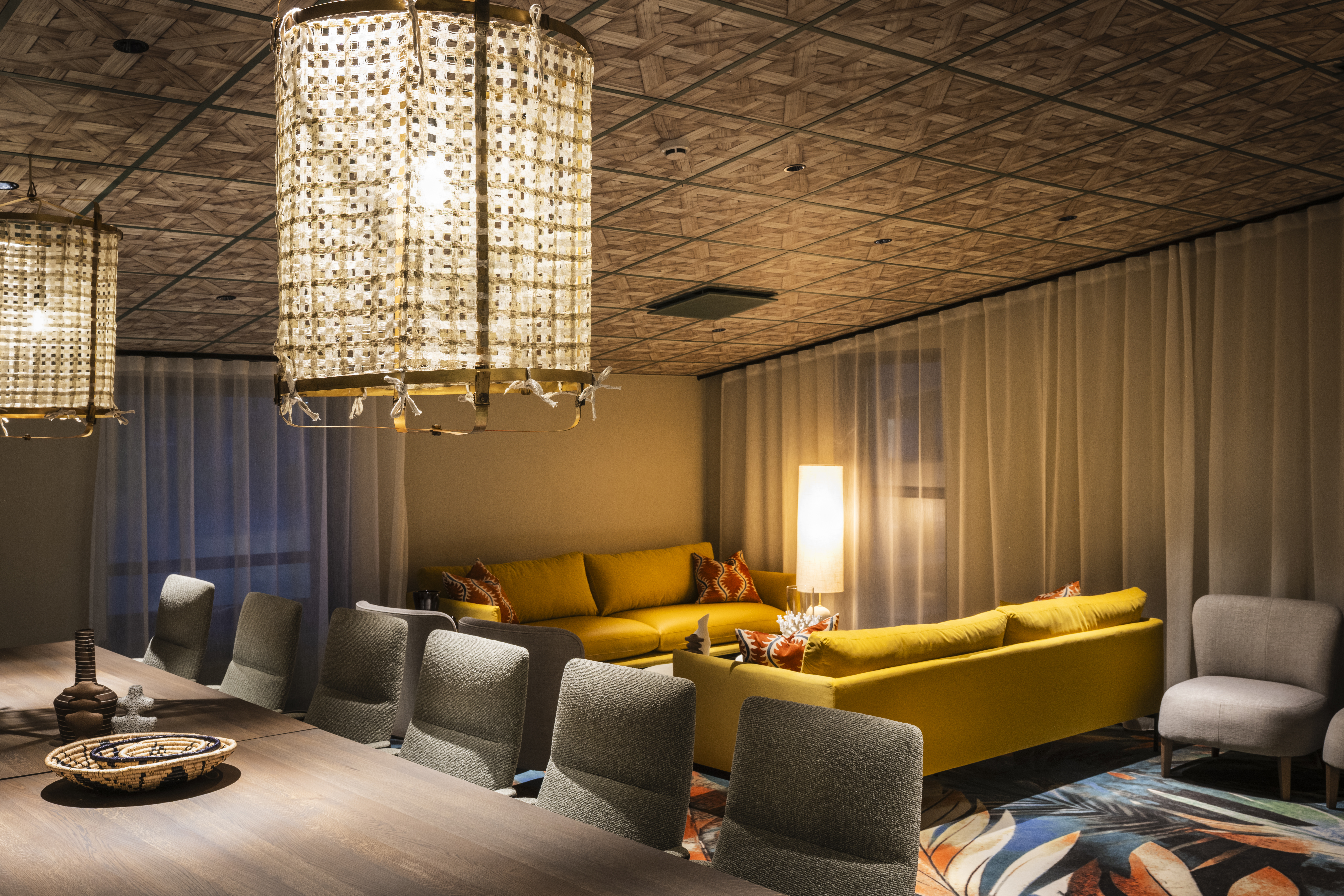 En stor, gul hjørnesofa er arrangeret rundt om en glødende lampe i et svagt oplyst rum, med mønstrede tæpper og komfortable stole omkring et træbord. Stoftaglamper hænger over.