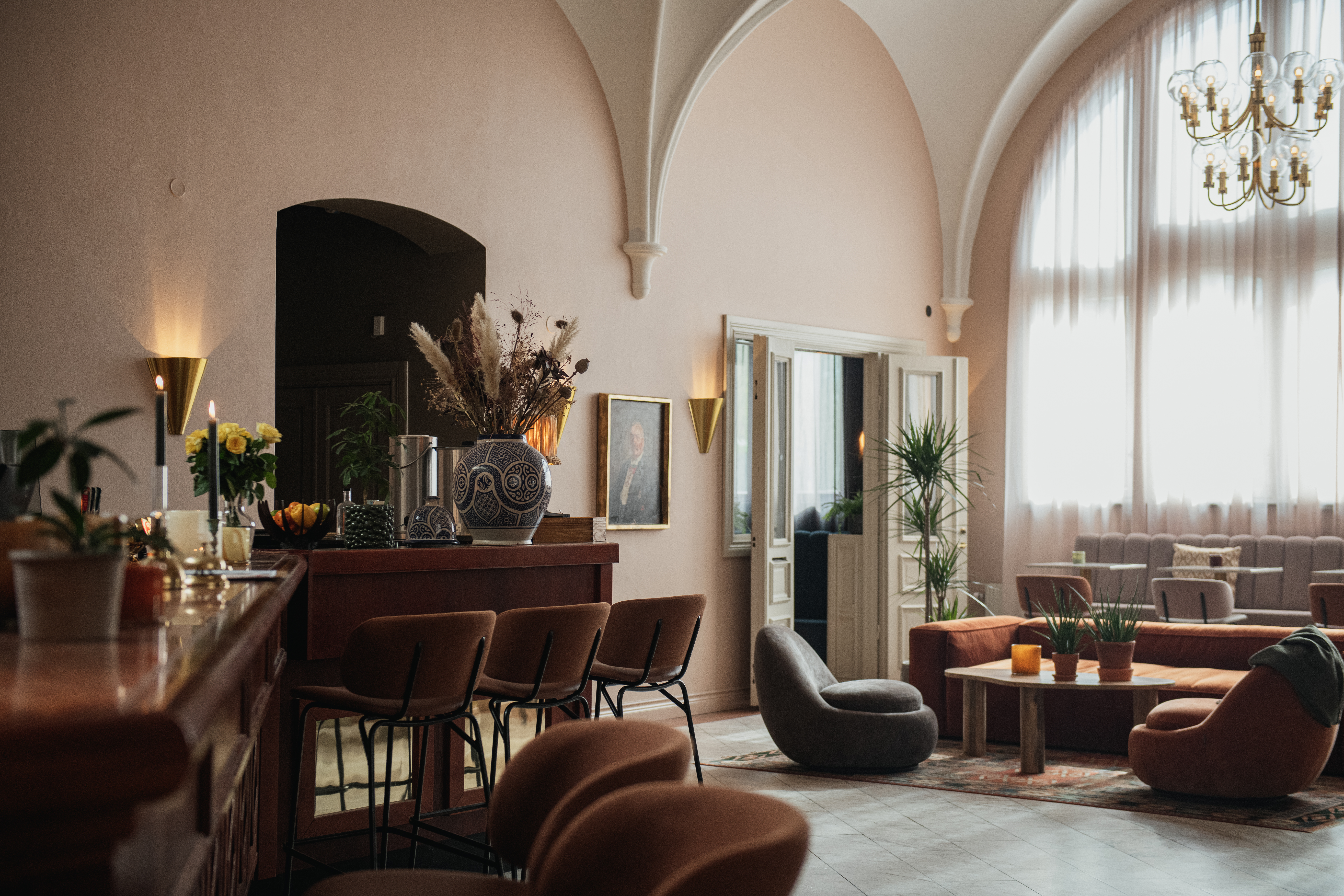 Bardisk med stole og dekorative genstande. Sollys strømmer ind gennem hvælvede vinduer og oplyser hyggelige siddeområder og potteplanter i en elegant, blidt oplyst lounge. Lysekrone hænger fra højt loft.
