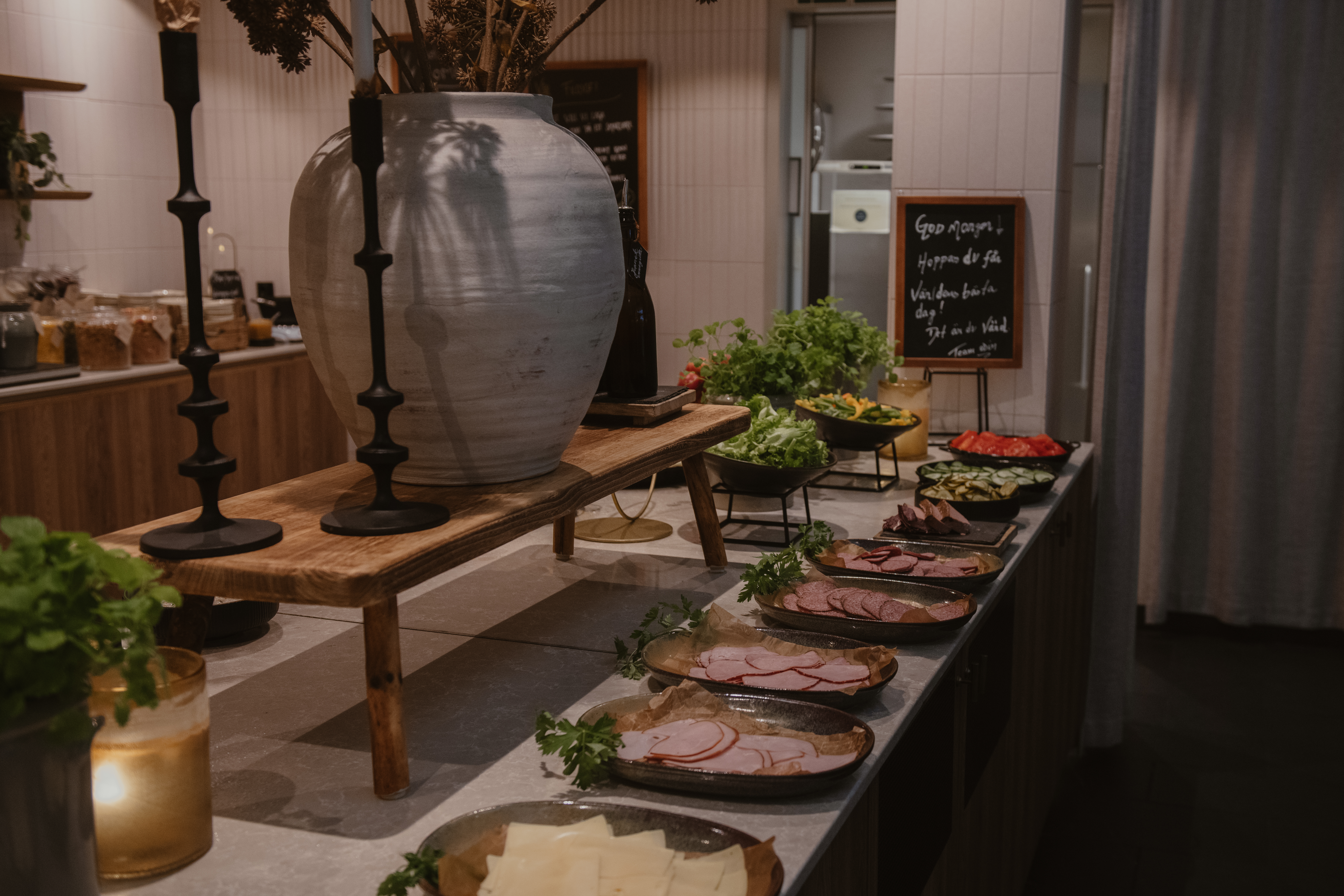 Buffébord med olika köttpålägg, grönsaker och örter. Stor vas och ljushållare pryder uppsättningen. En griffeltavla i bakgrunden lyder: "Gør morgen maden der hjemme værd at stå op for."