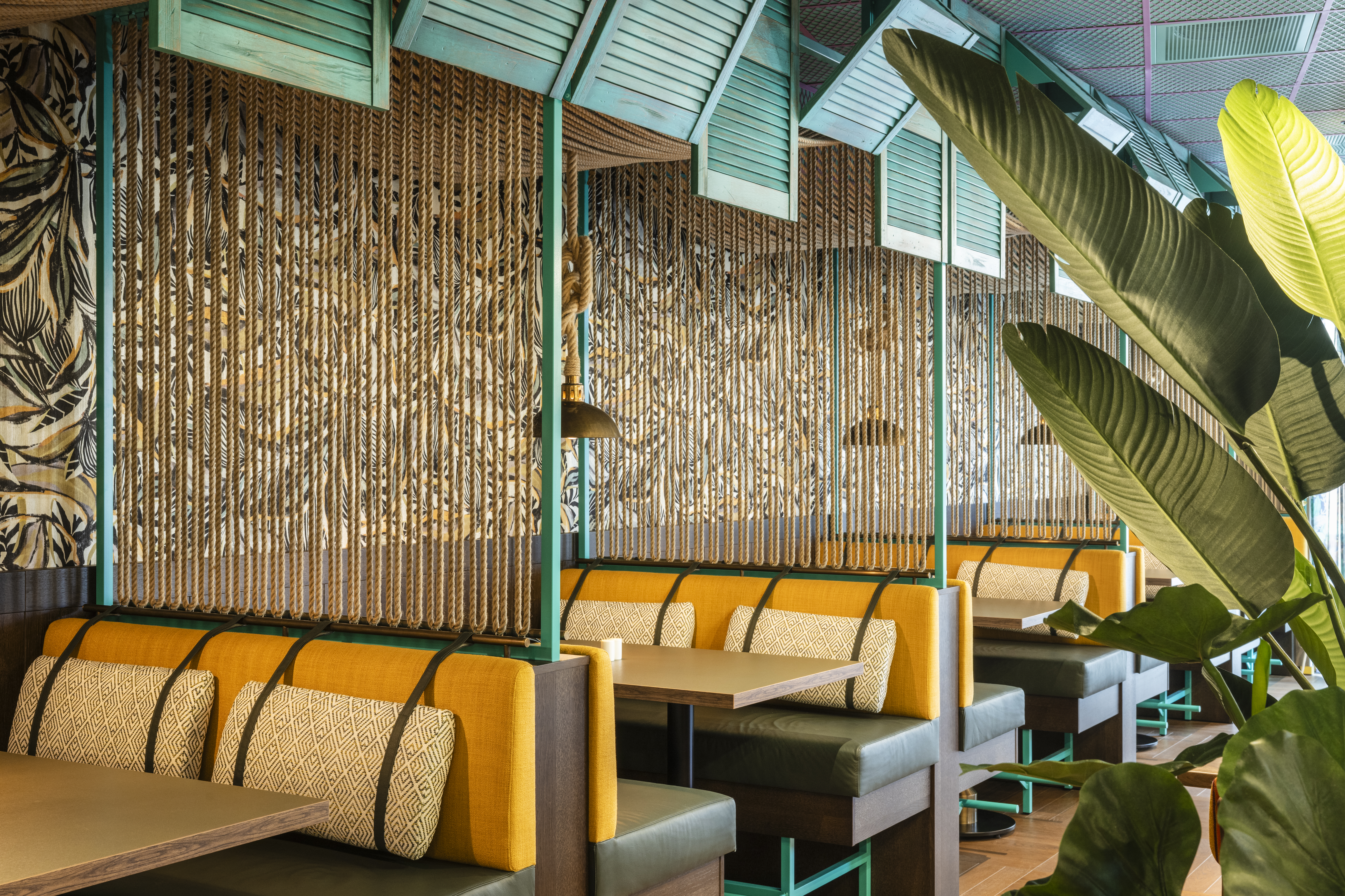 Farverige båse med gule puder og mønstrede puder arrangeret i en restaurant. Rummet har rebopdelere, tropisk planteindretning og teal farvede loftspaneler, der skaber en livlig, hyggelig atmosfære.