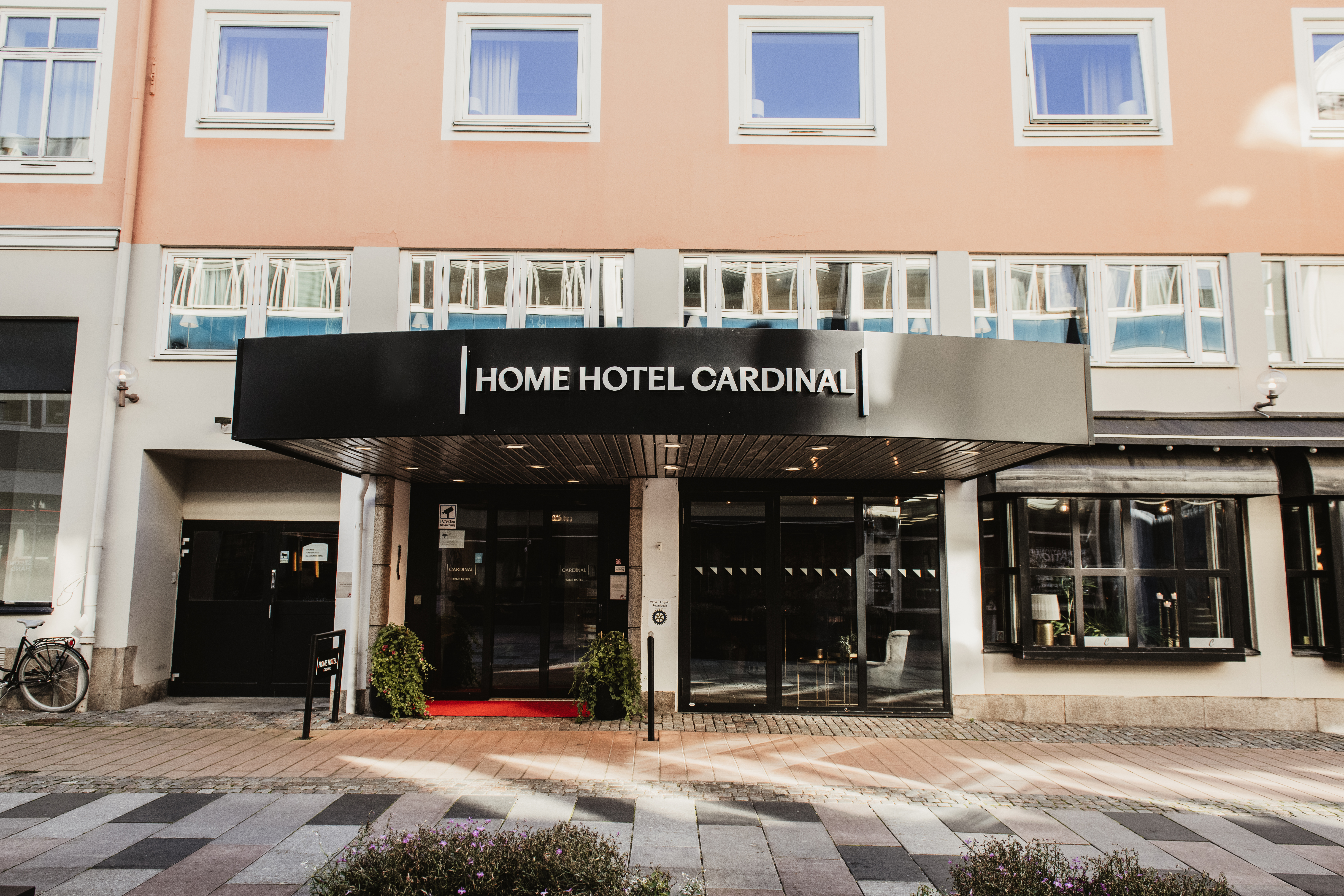 Kutsuva hotellin sisäänkäynti, jossa kyltti HOME HOTEL CARDINAL. Punainen matto ja ruukkukasvit koristavat sisäänkäyntiä.