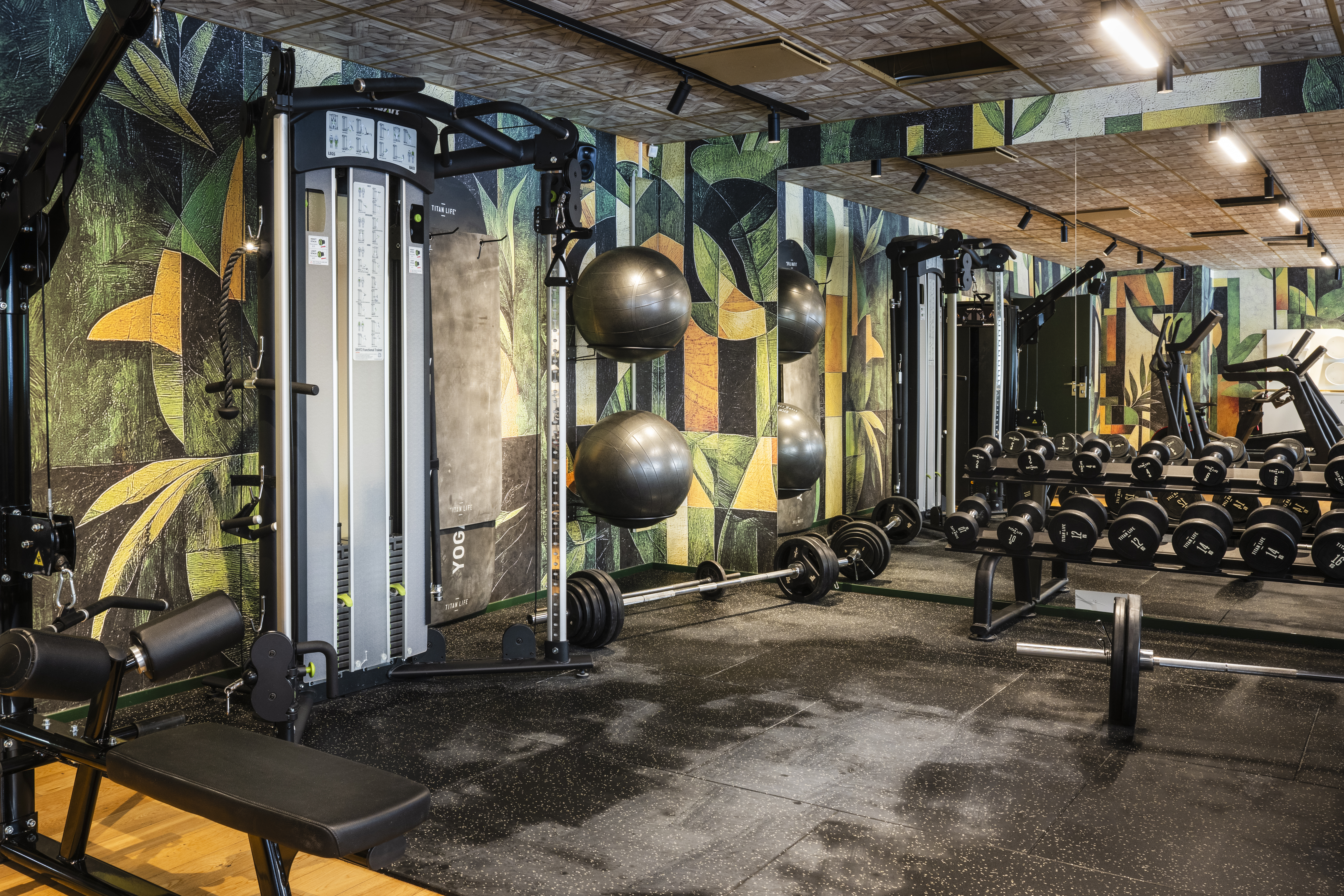 Fitnessudstyr, placeret i et moderne fitnesscenter, inkluderer håndvægte på stativer, træningsmaskiner og træningsbolde mod en farverig geometrisk-mønstret væg, der skaber en energisk træningsstemning.
