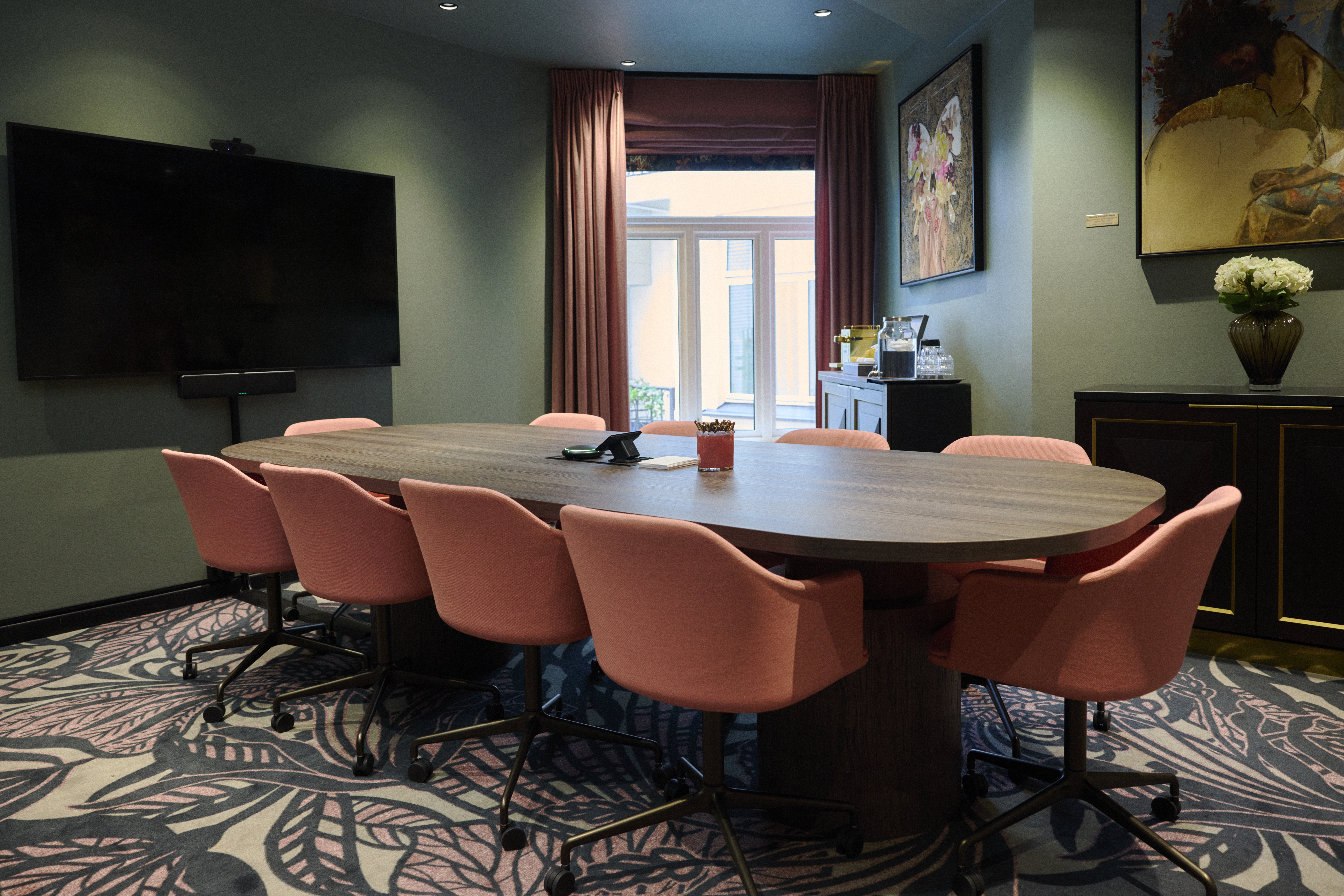 Conference table surrounded by pink chairs, set in a room with a large screen, floral carpet, and artwork. Light filters through windowed curtains, softening the modern decor.