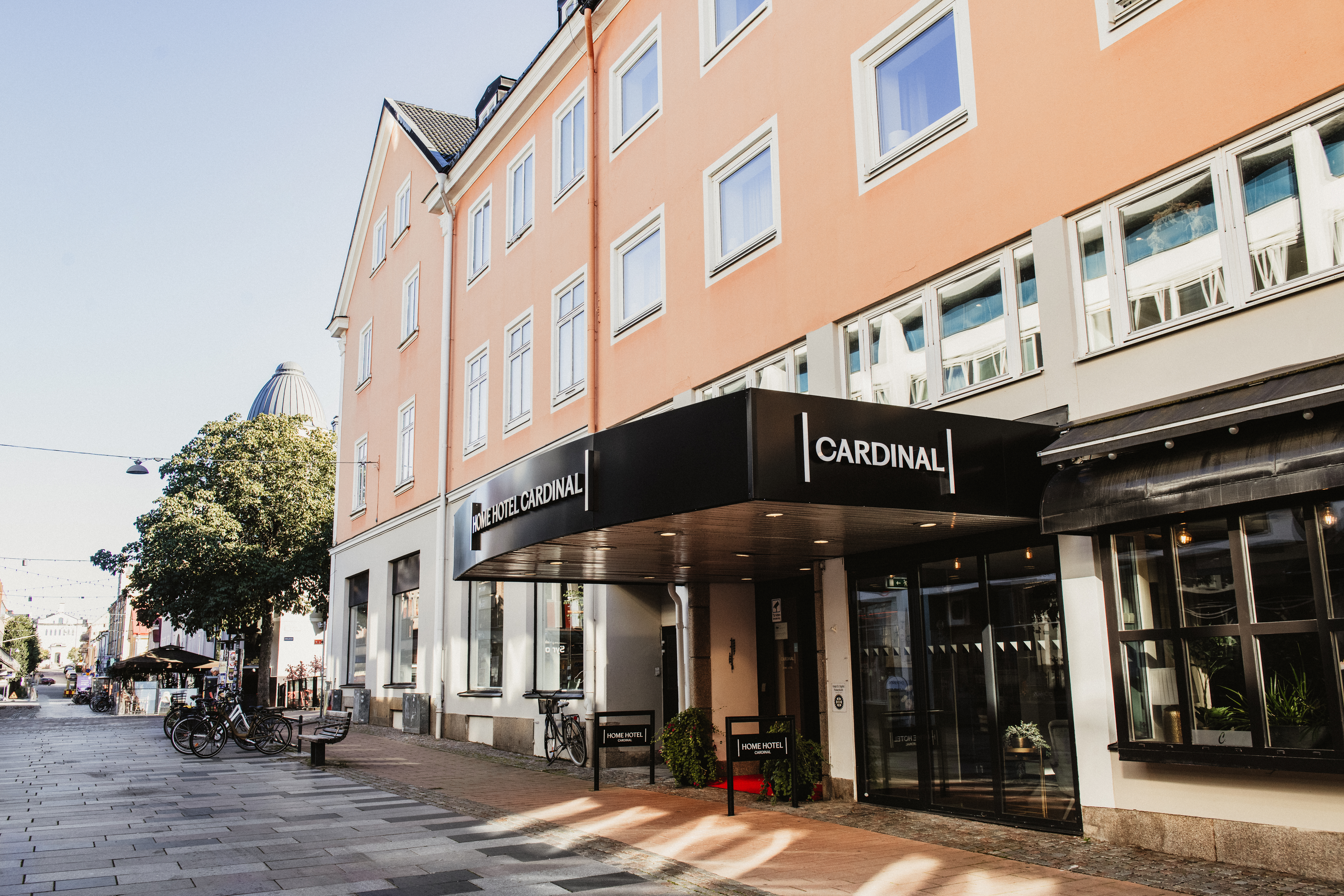 Hotellin sisäänkäynti aurinkoisella kadulla, jossa on musta markiisi, jossa lukee HOTEL CARDINAL. Polkupyöriä on pysäköity lähelle.