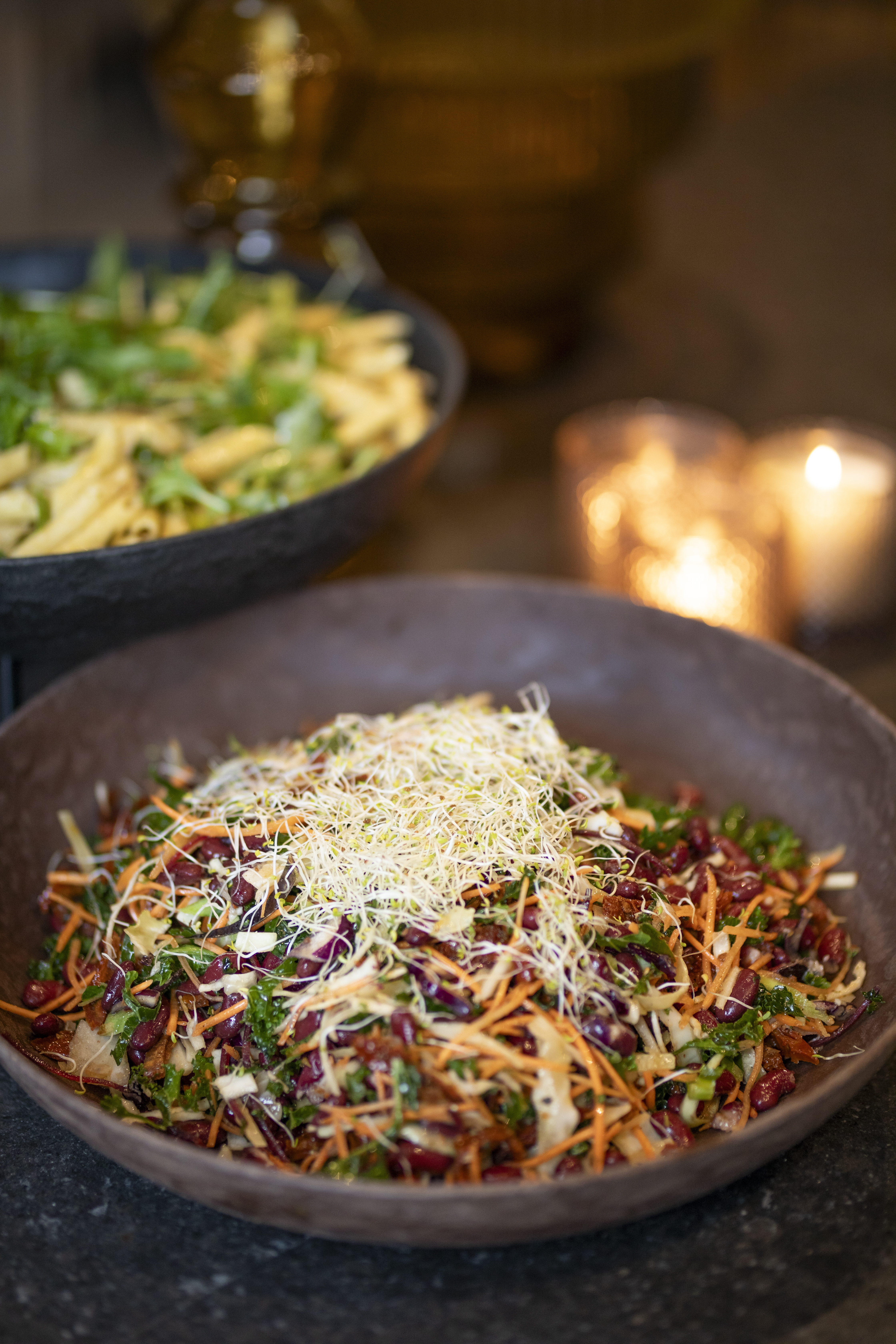 A colorful salad with shredded vegetables and sprouts sits in a brown bowl. Another bowl with pasta and greens in the background. The setting is warmly lit by candles.