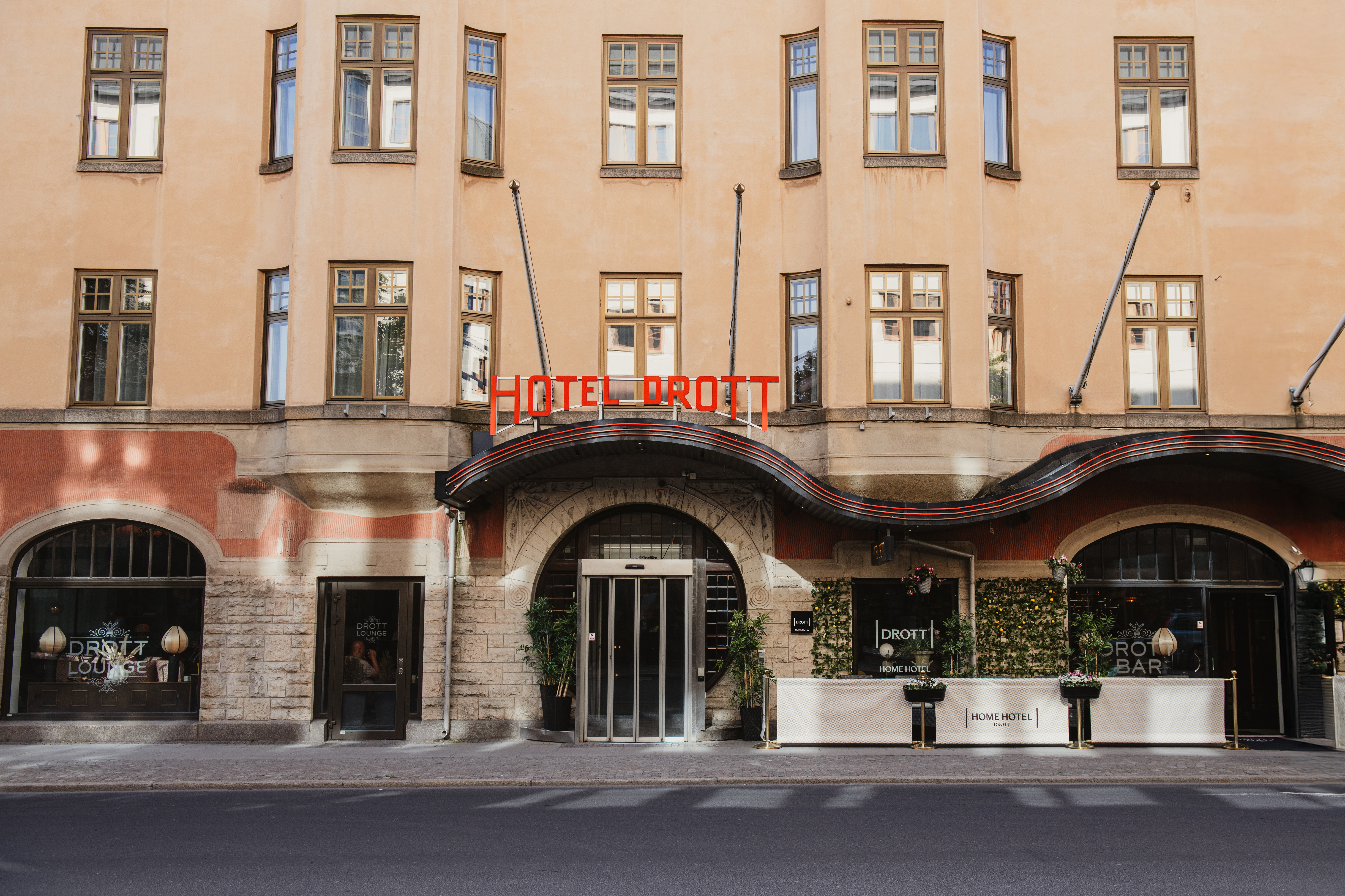 Byggnadsfasad visar "HOTEL DROTT" i röd neon, med välvda ingången omgiven av växter. Området inkluderar fönster och skyltar för "DROTT LOUNGE" och "DROTT BAR".