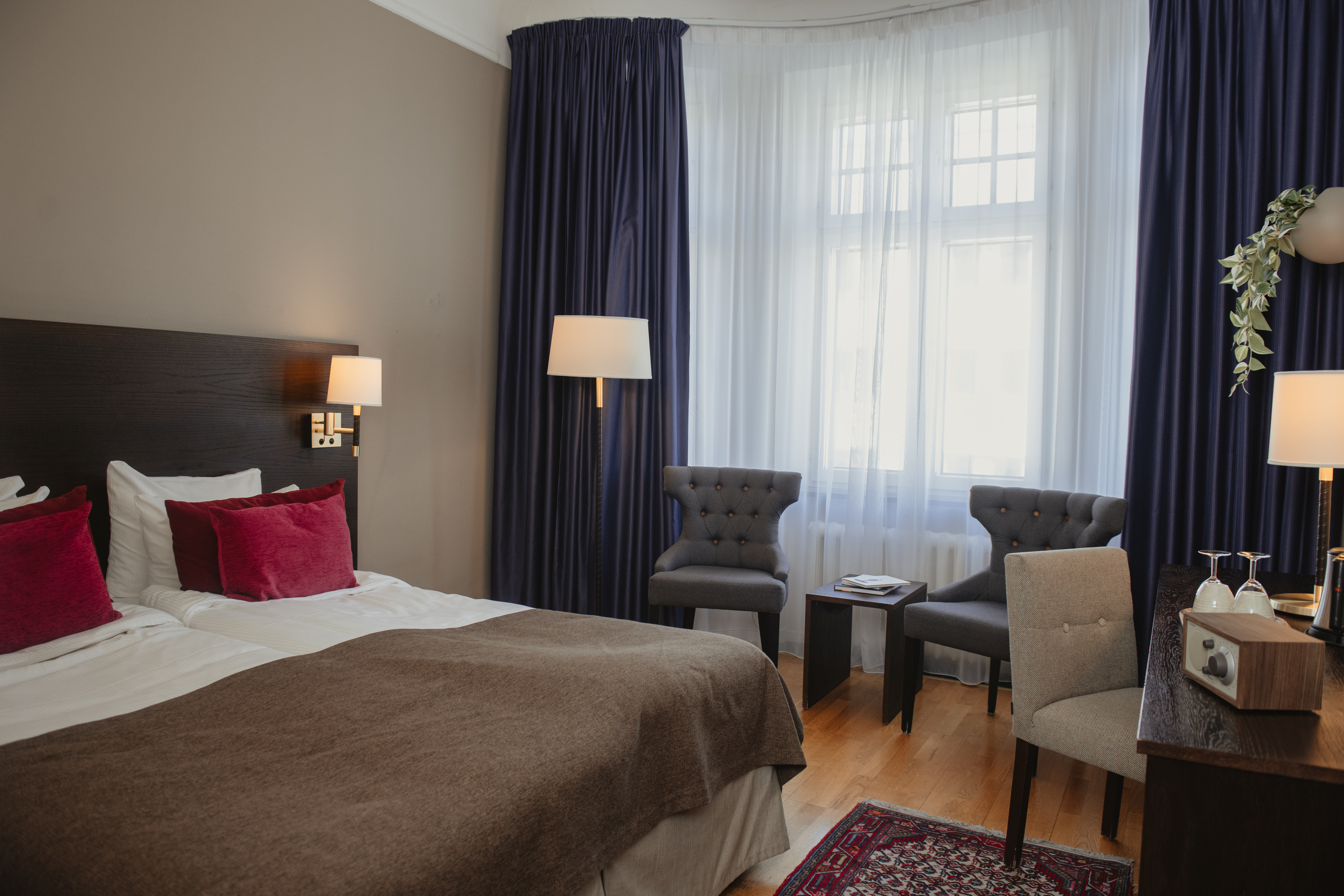 Dubbelsäng med brun bädd och röda kuddar i hotellrum. Två grå stolar vetter mot ett litet bord vid ett fönster med blå gardiner. Ett skrivbord har en lampa och dekorationer. 