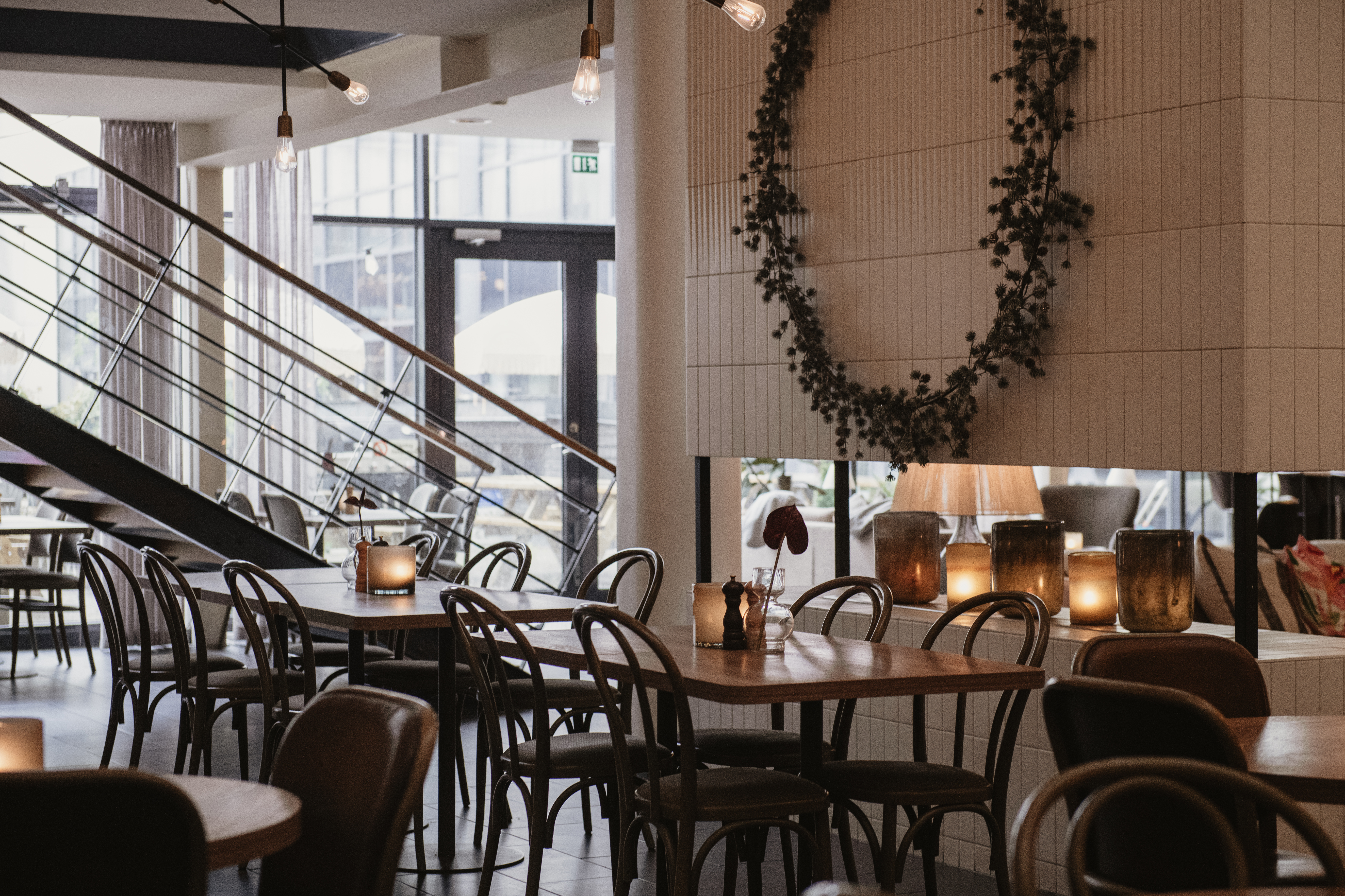 Bord och stolar fyller ett mysigt, svagt upplyst kafé med varma lampor och dekorativa växter på väggen, trappa leder upp och stora fönster släpper in naturligt ljus.