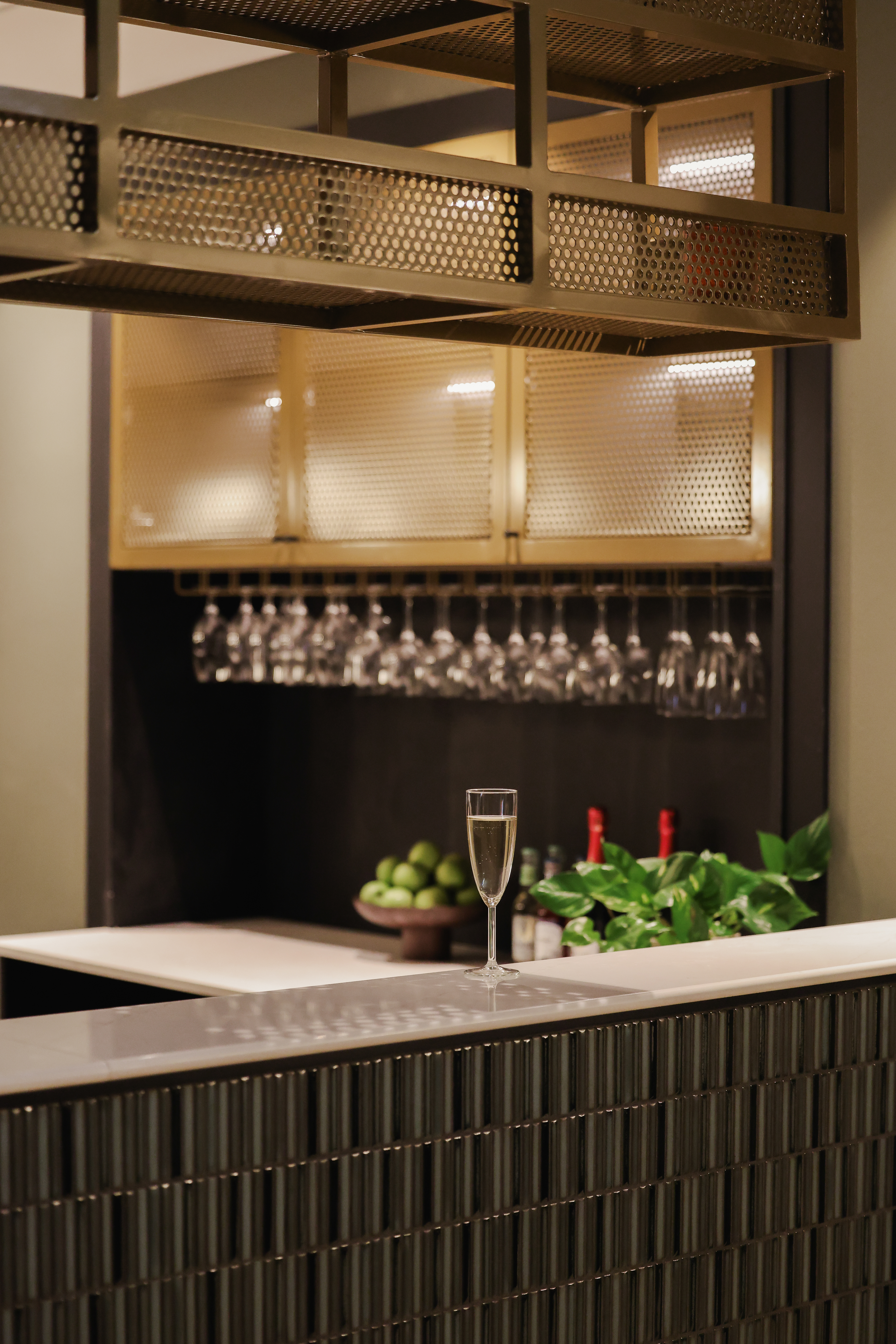 Ett glas champagne står på en modern bardisk. Hängande glas och flaskor är snyggt arrangerade på hyllor, omgivna av växter och frukt, vilket skapar en elegant atmosfär.