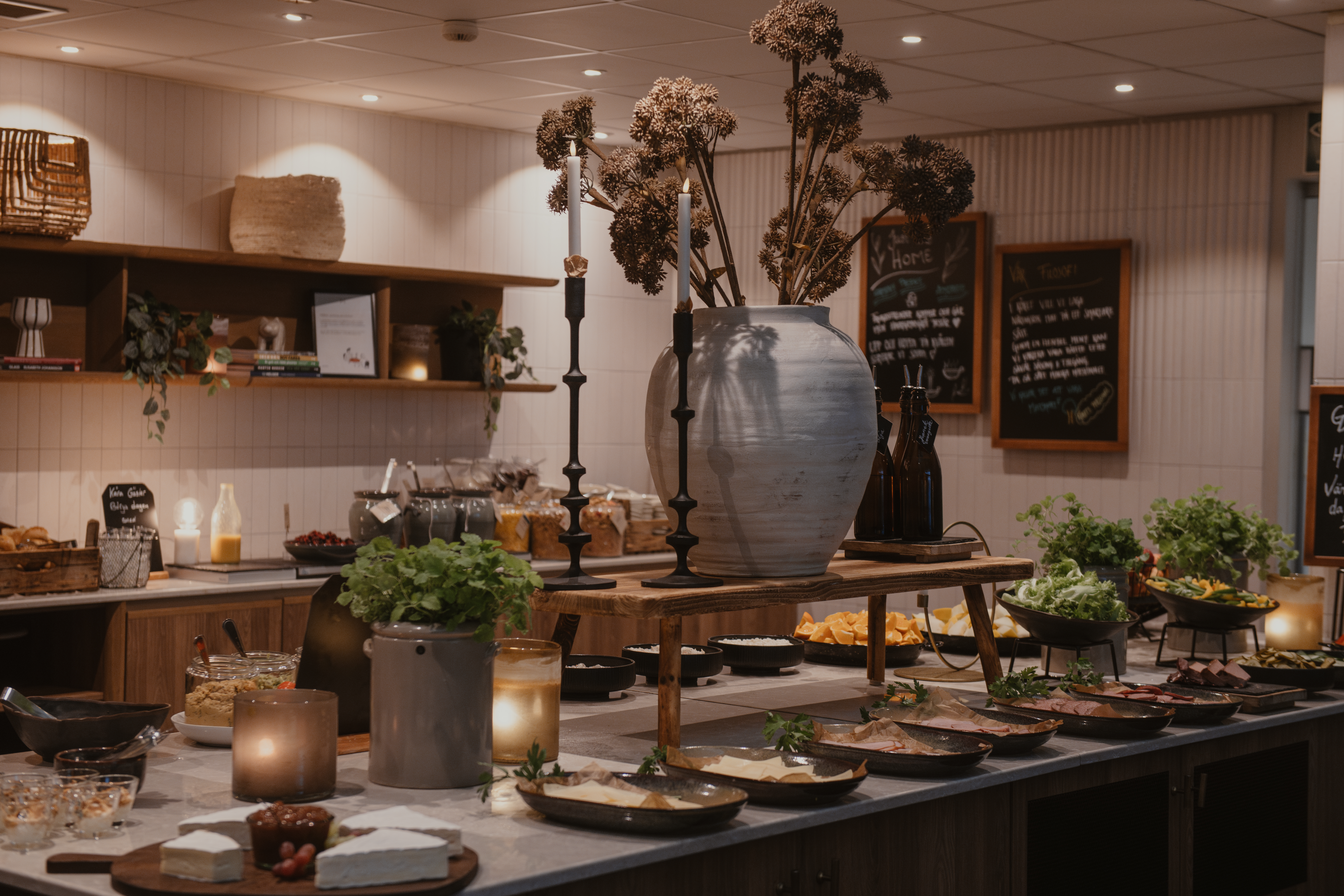 En buffébord visar maträtter och ljus i ett varmt upplyst rum. Menytavlor med texten "Menu," "Glutenfri," och "Vegan" syns på väggarna bakom arrangemanget.