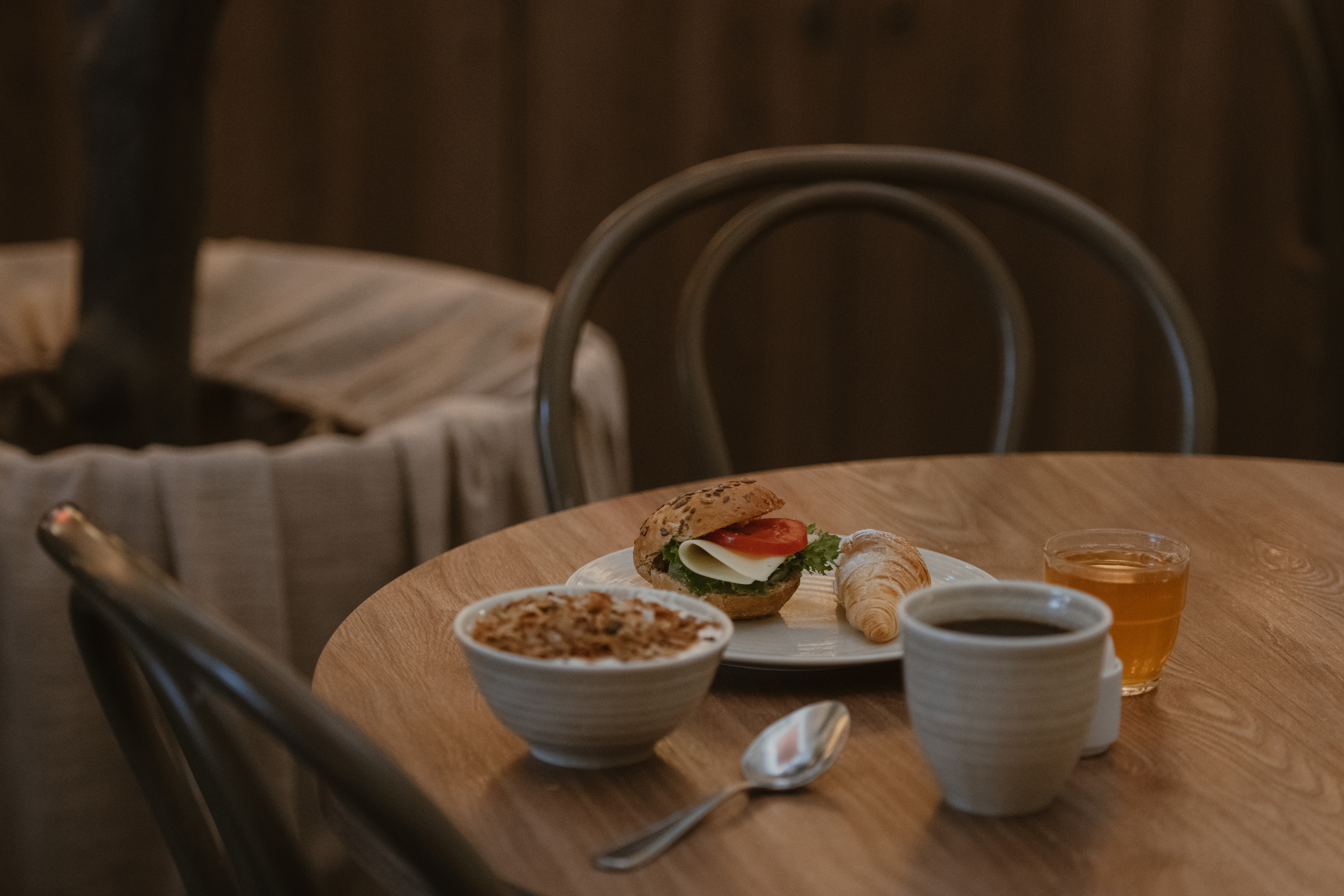 Smörgås med grönsaker och kalkon ligger på en tallrik bredvid en kaffekopp, croissant, skål med flingor och juiceglas. Placeras på ett runt träbord med stolar i en mysig miljö.