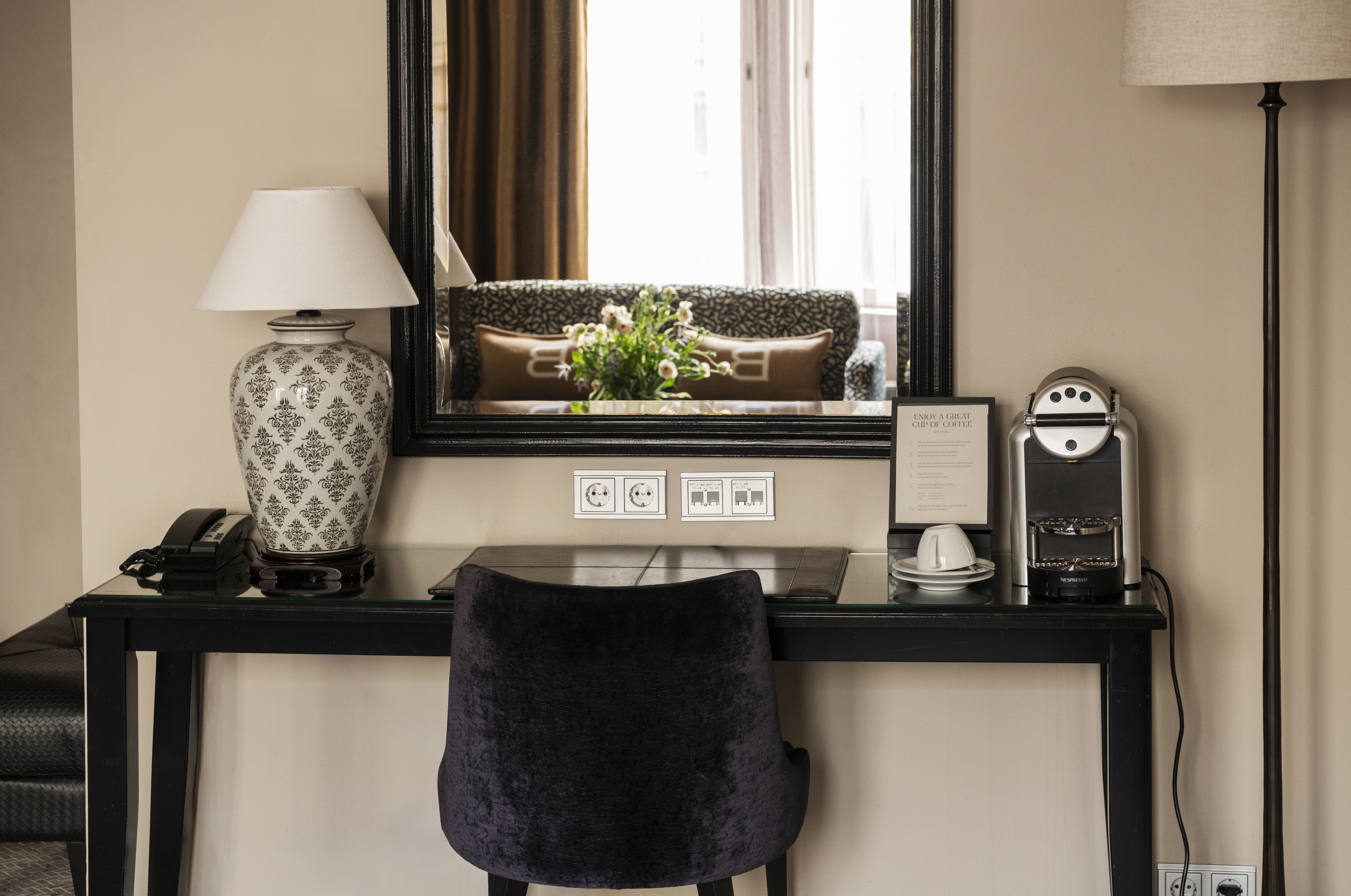 A desk with a black chair and a lamp on the left; a coffee machine and framed picture on the right. Large mirror reflects a sofa and bouquet, creating a cozy atmosphere.