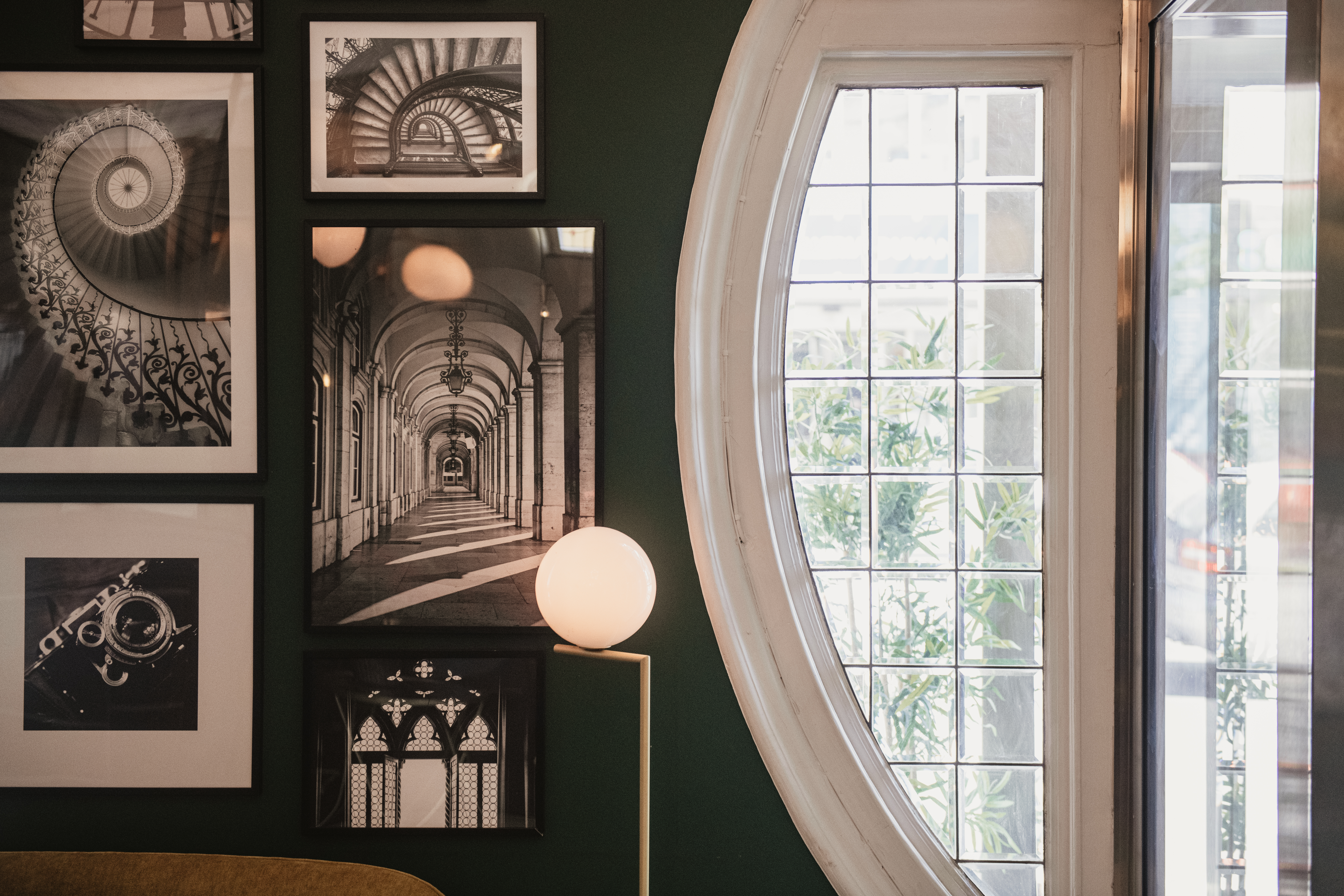 Flera inramade svartvita arkitekturfotografier hänger på en mörk vägg, upplysta av en sfärisk lampa; ett ovalt fönster med rutnät skymtar grönska utanför.