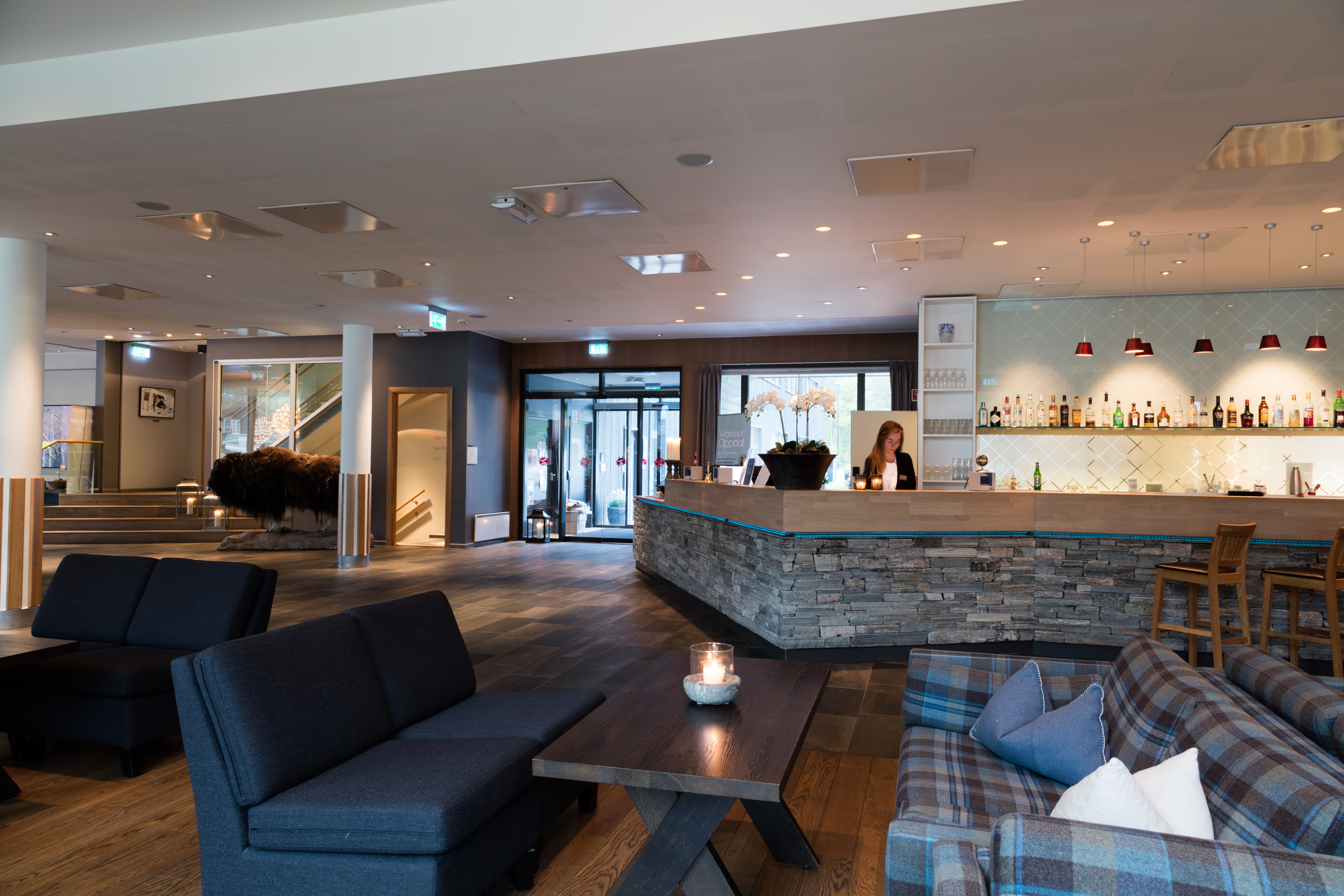 Resepsjonsdisk med steinfront og en bartender som forbereder drinker, plassert i en moderne salong med komfortable sitteplasser, tregulv og stemningsbelysning.