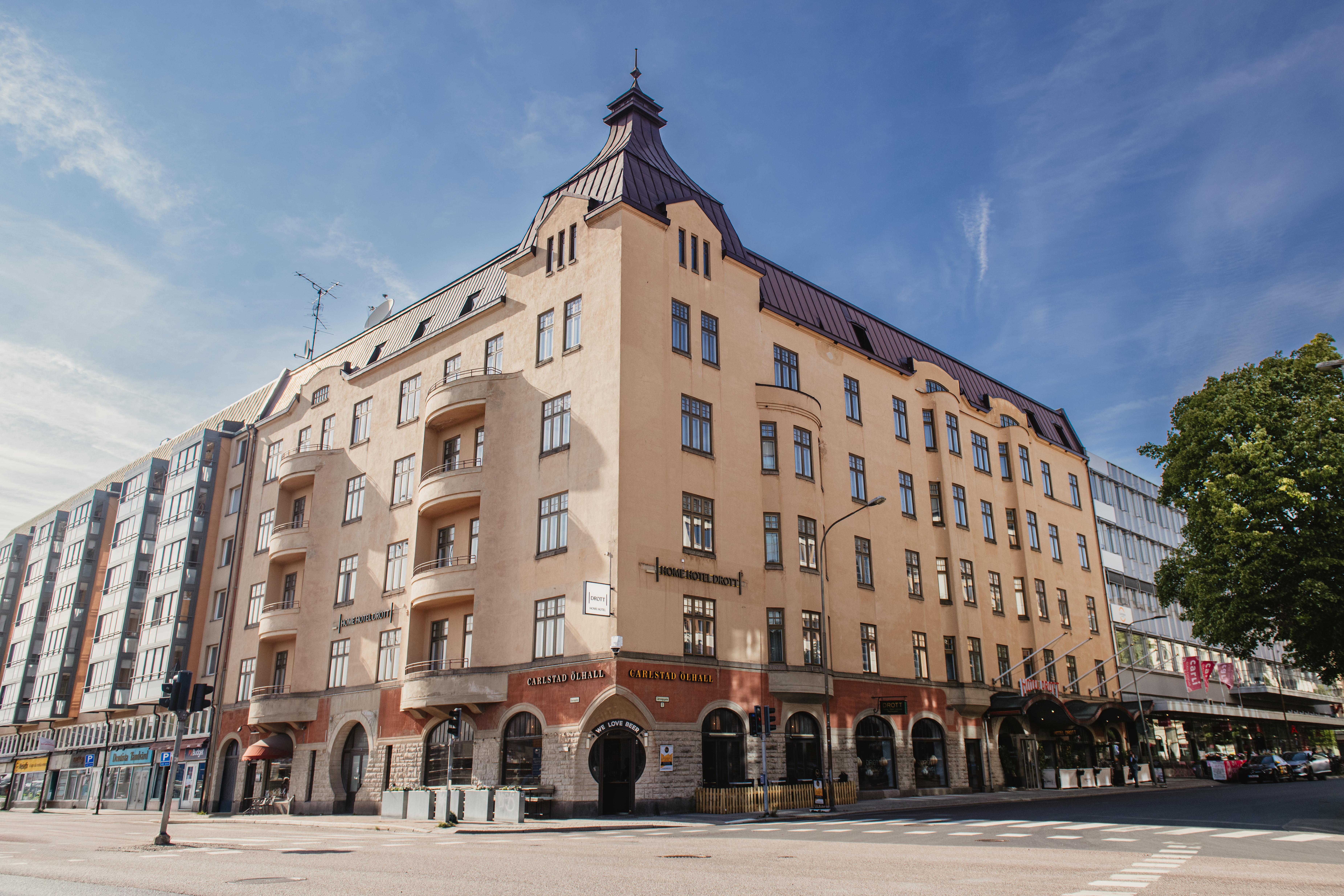 Ett beige fler-våningshus står på ett gatuhörn med välvda fönster, balkonger och brant tak. Ett träd och andra byggnader omger det. Texten lyder "Fennia Salonki" och "Fennia Restaurant".