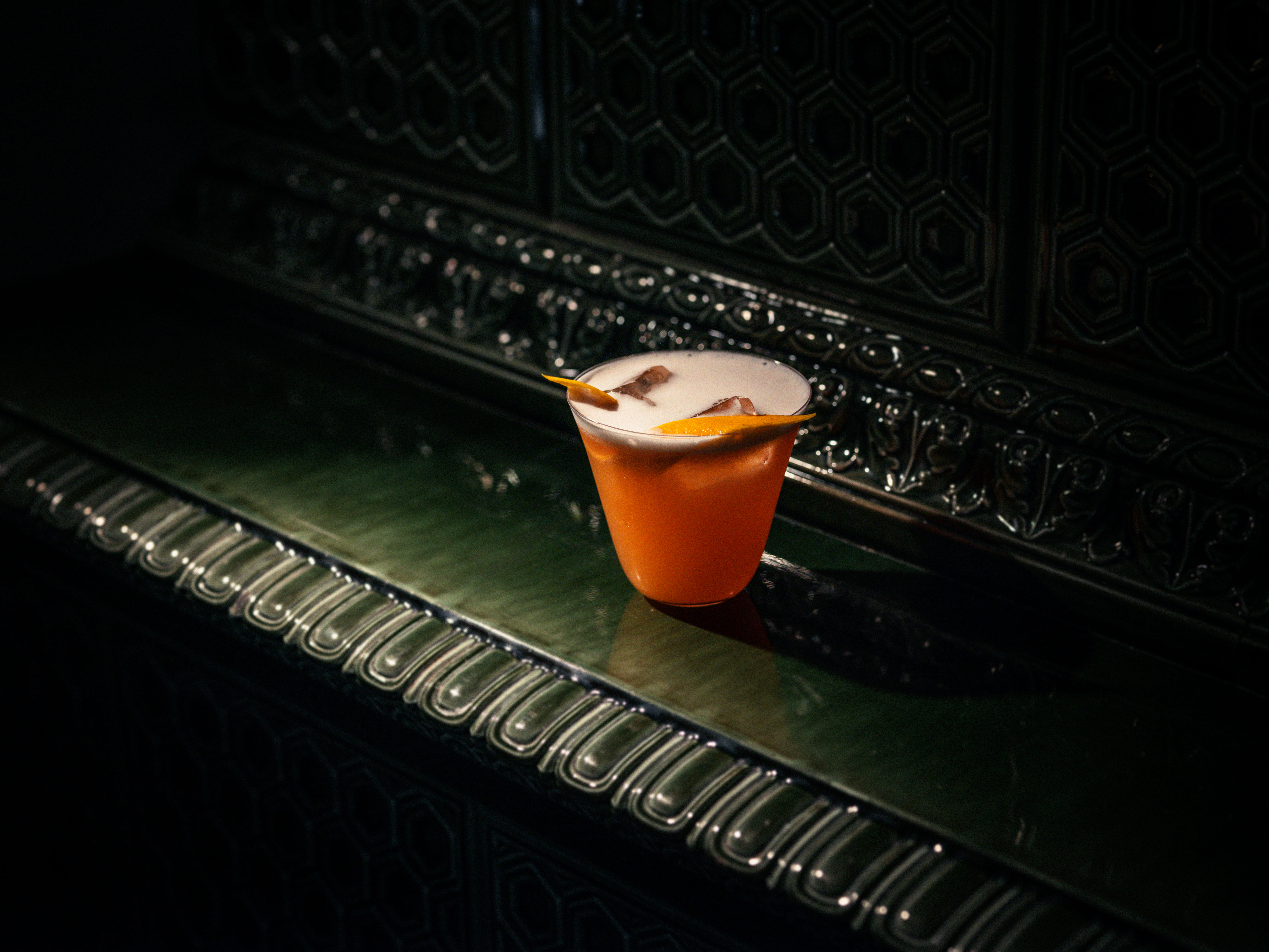 A vibrant cocktail with foam and an orange garnish, set against a dark, ornate background at Hotel Kämp – Kämp Bar.