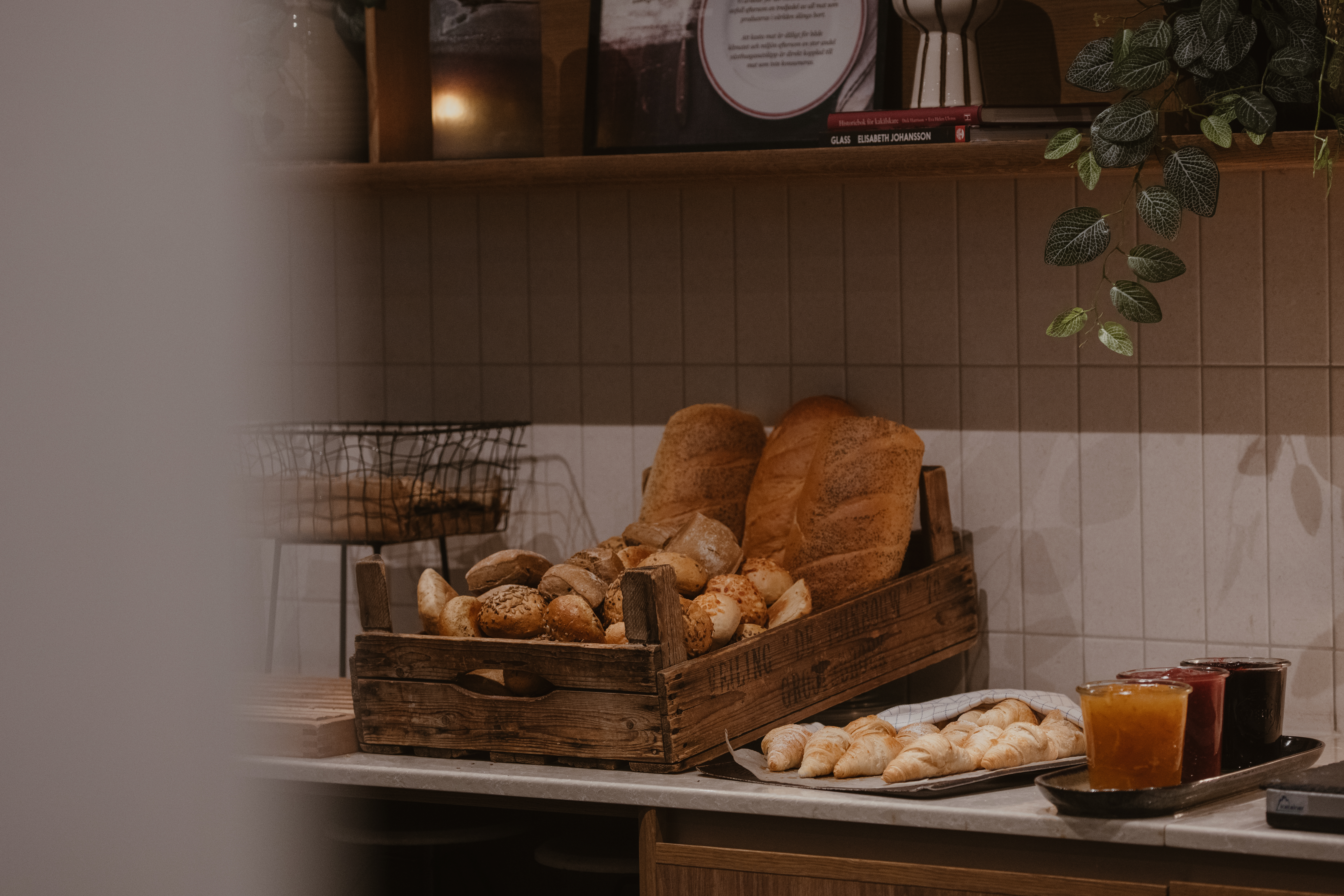 Ett trälåda fylld med blandat bröd står på en köksbänk. Burkar med apelsin och röd sylt står bredvid, under en hylla med dekorativa växter.